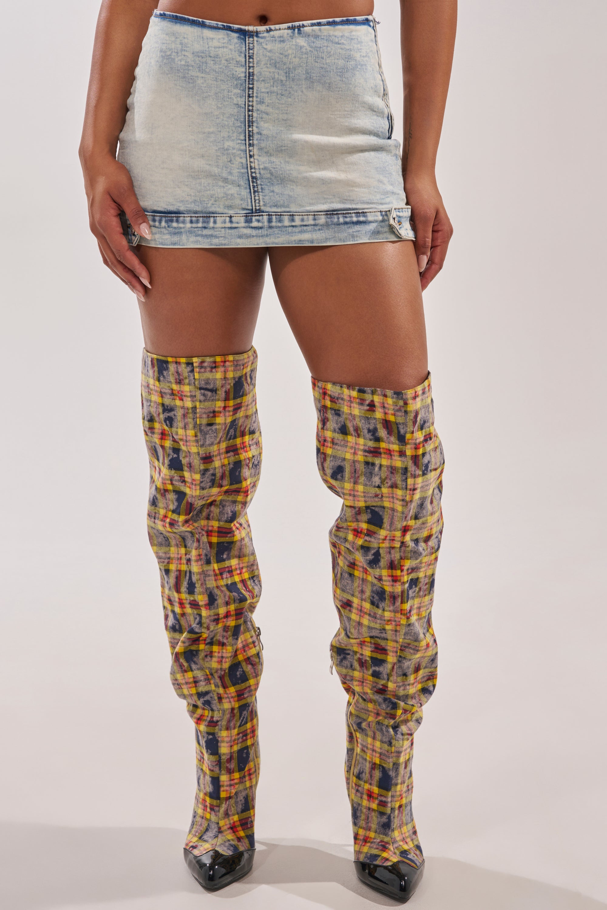 Wearing the BABY GIRL DENIM MINI SKORT in light blue with a high-rise waist, paired with tall, slouchy plaid boots featuring pointed black toes, this look channels a cool Y2K vibe against a plain background. 1