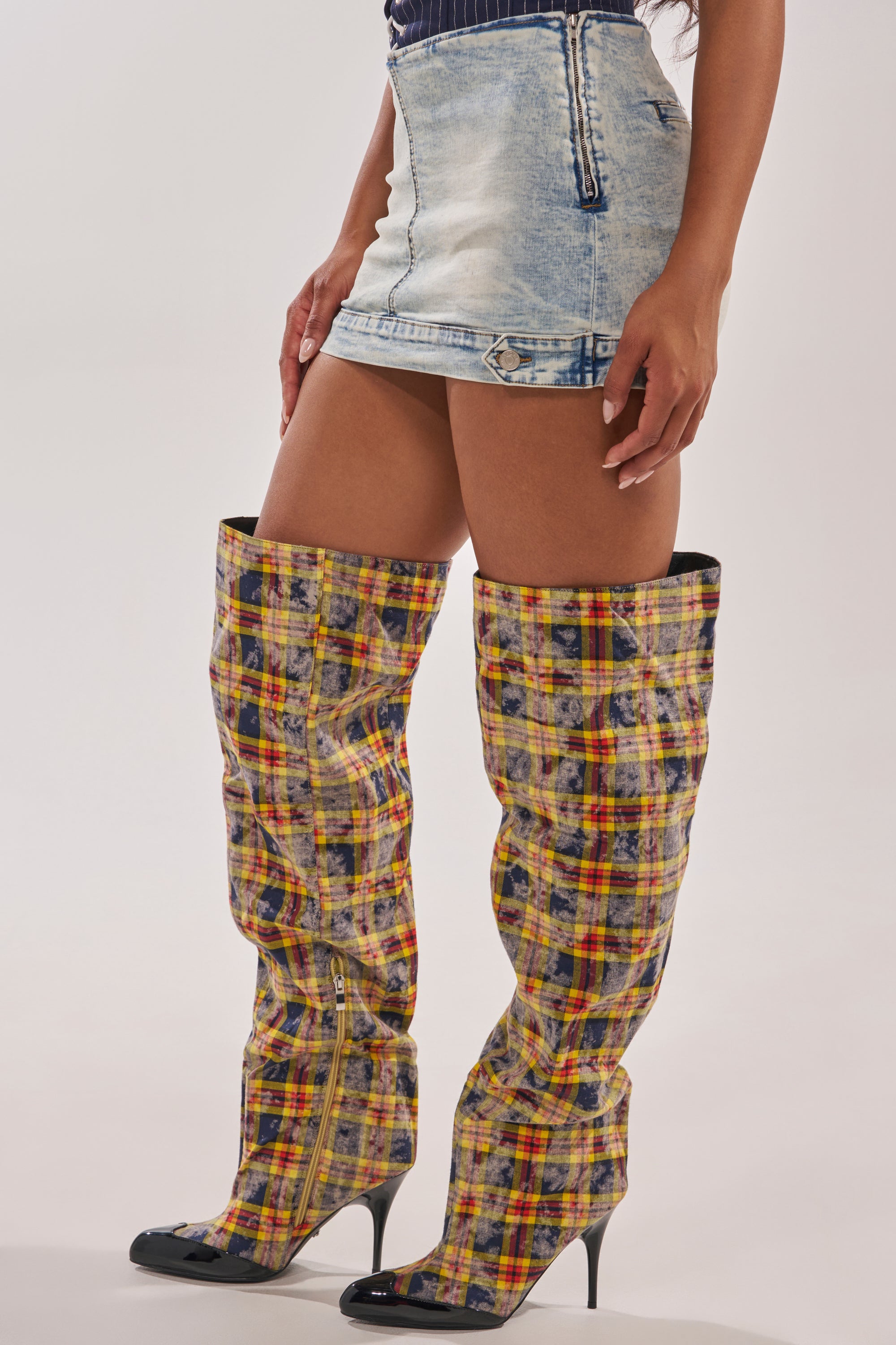 A person showcases the BABY GIRL DENIM MINI SKORT in light blue, styled with a high-rise waist and paired with tall, slouchy plaid boots with pointed black toes and high heels, creating a trendy Y2K look against a plain light backdrop. 3