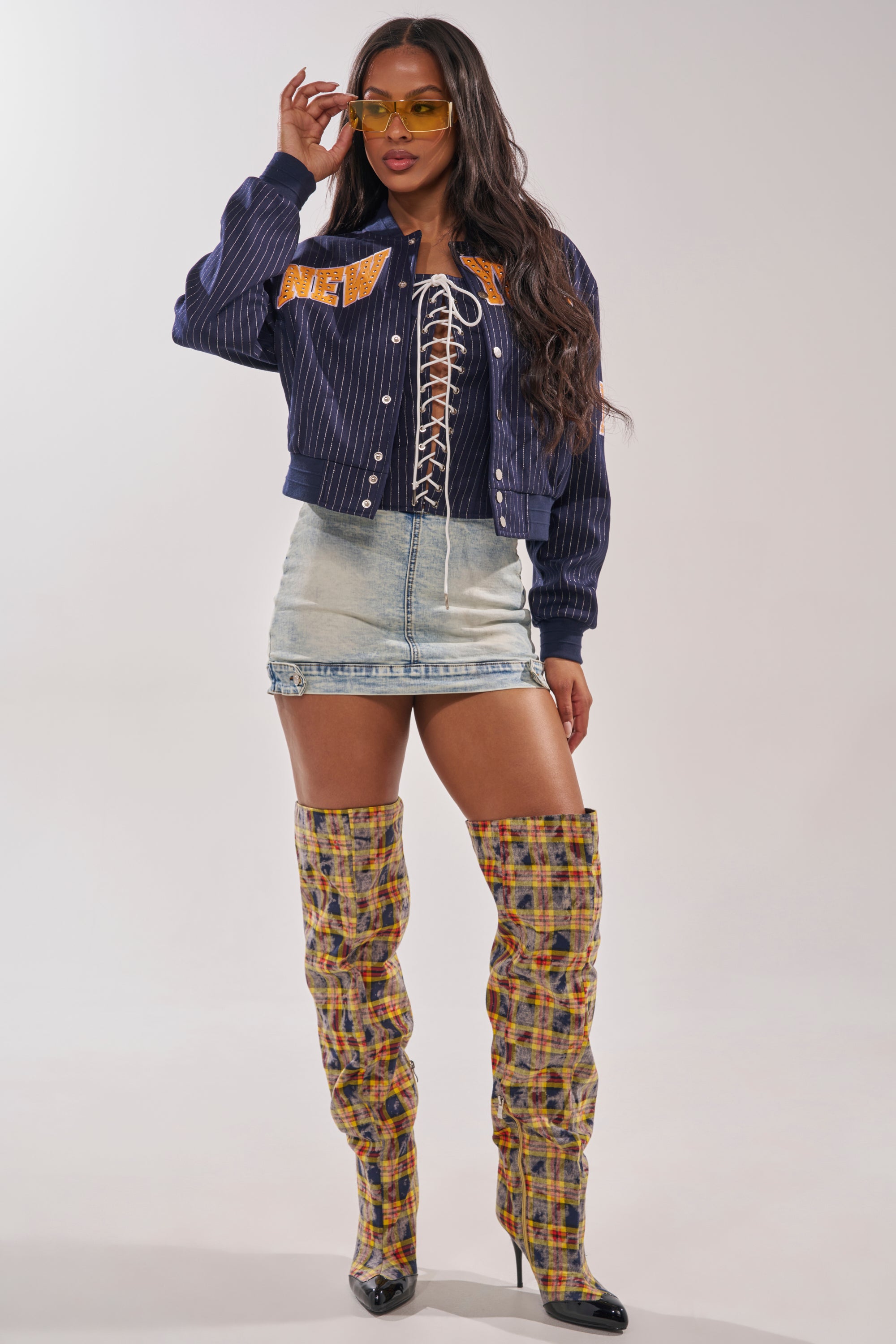 Woman poses in BABY GIRL DENIM MINI SKORT, a navy varsity jacket, black lace-up top, tall plaid pointed-toe boots, and yellow sunglasses. She has long wavy hair and stands against a plain light background. 7