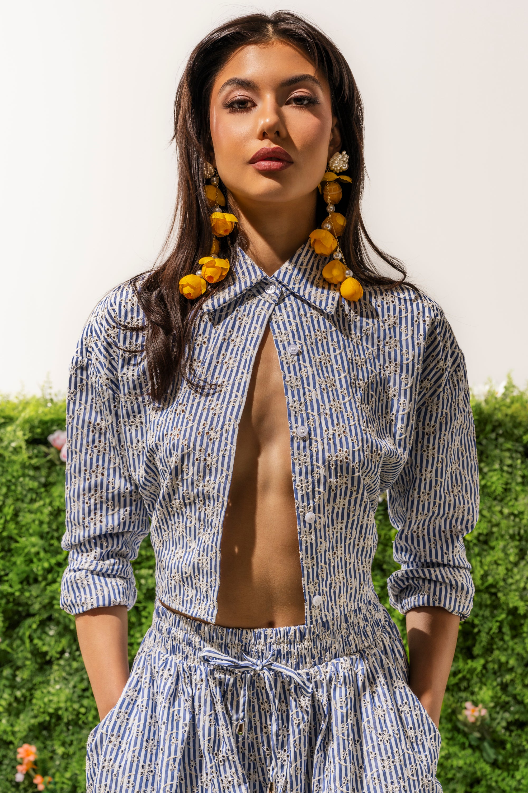 A woman with long dark hair and yellow-accent earrings stands outdoors in the BLOSSOM CROP BUTTON DOWN, a blue and white patterned top with floral embroidery, paired with matching pants. She looks confident against a backdrop of green foliage. 1