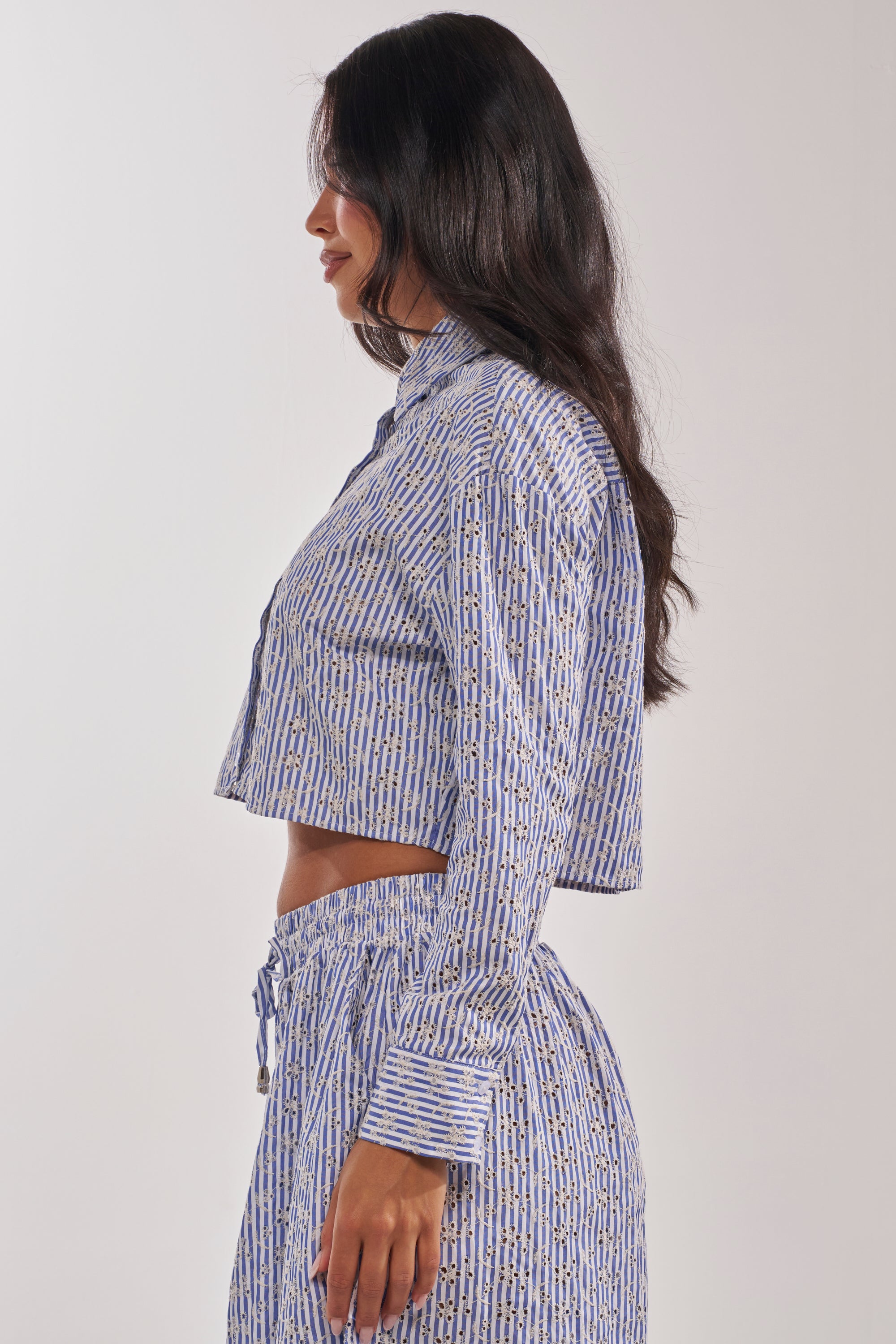 A woman poses in profile in the BLOSSOM CROP BUTTON DOWN—a blue and white striped cropped shirt paired with matching high-waisted pants. She has long dark hair and a neutral expression, set against a plain light background. 4