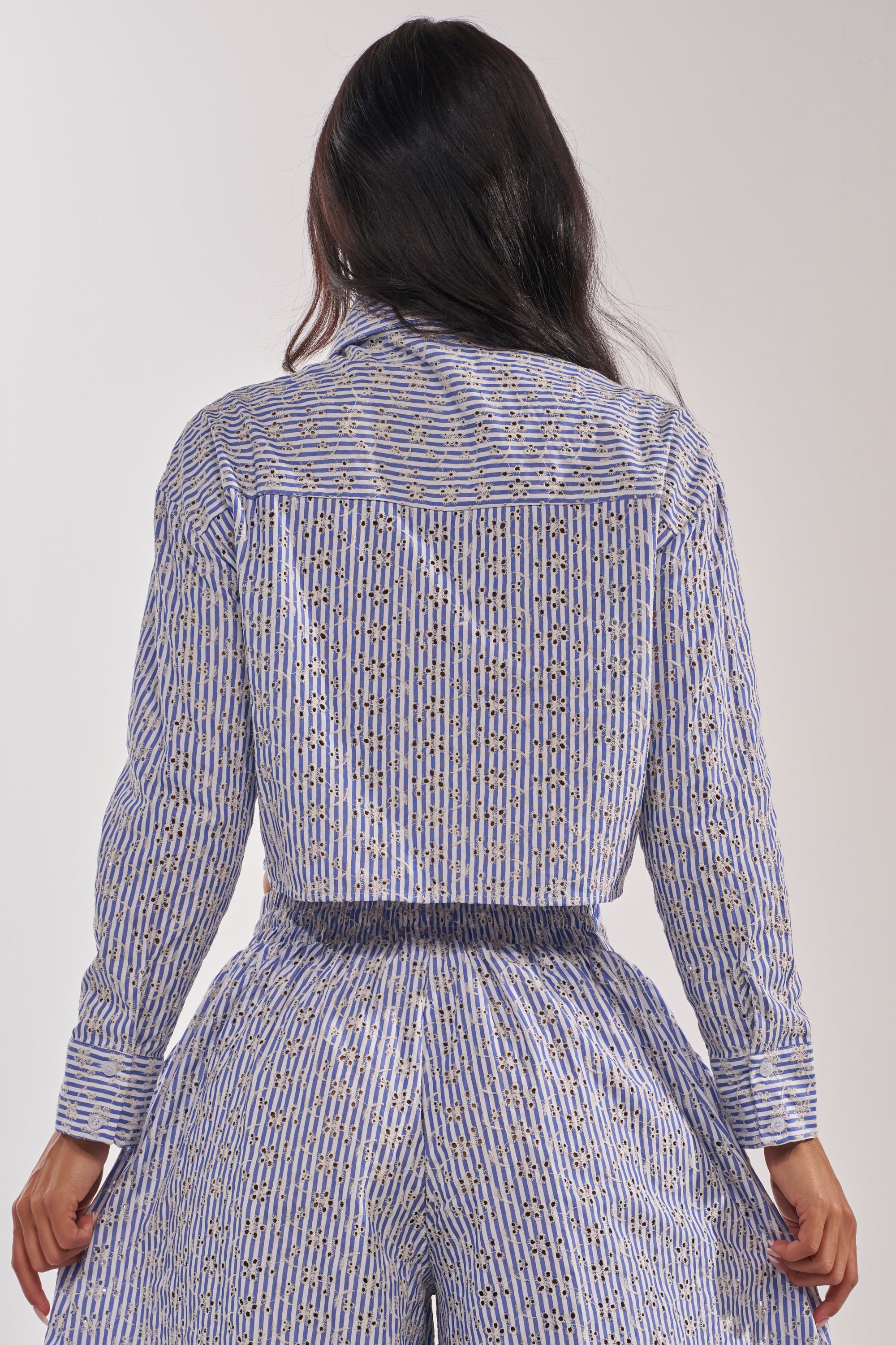 A person stands facing away, wearing the BLOSSOM CROP BUTTON DOWN—a blue and white striped top paired with high-waisted pants. Their long dark hair contrasts beautifully with the bold geometric print and yellow accents on the coordinated outfit. 5