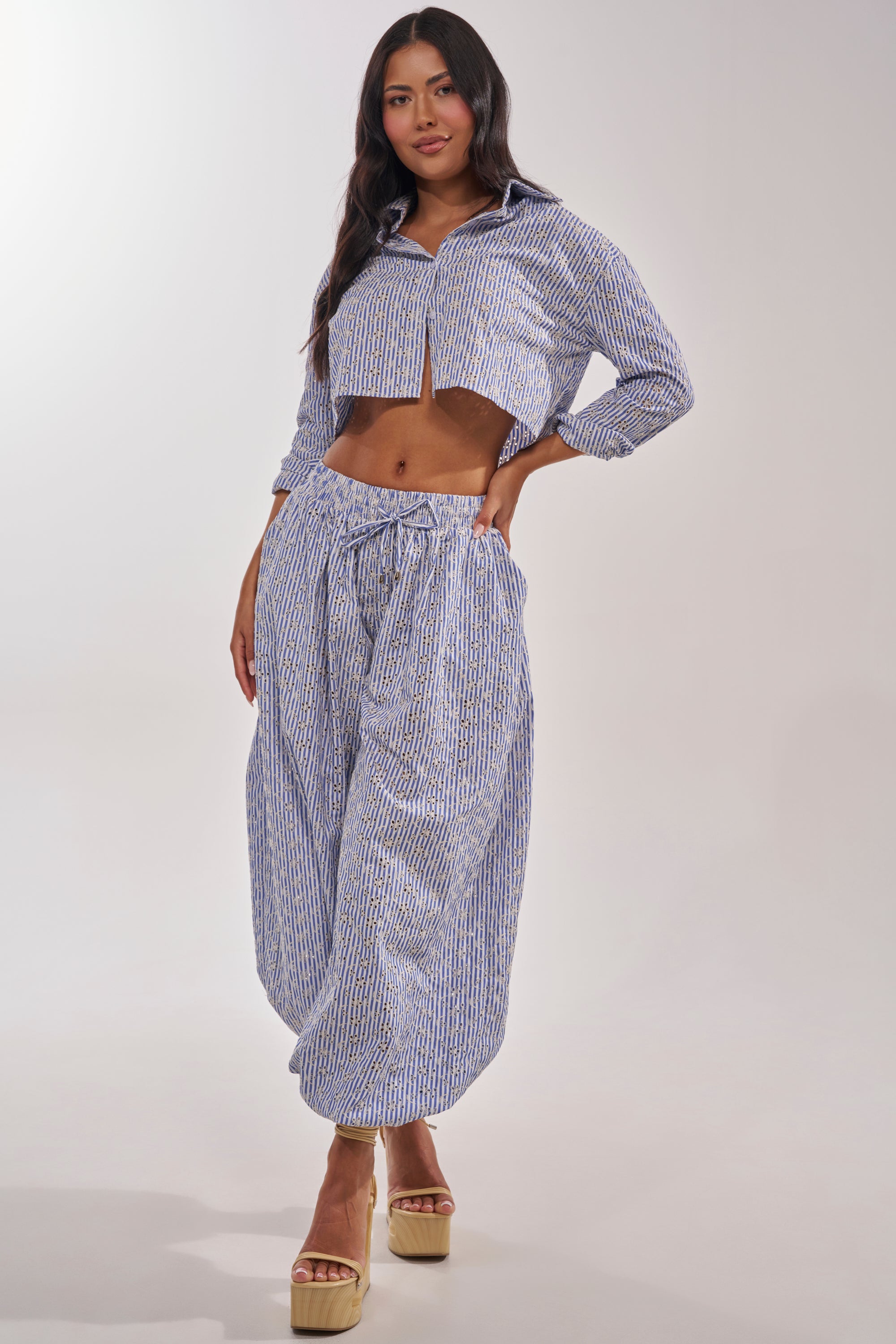 A woman poses in the BLOSSOM CROP BUTTON DOWN, a blue and white striped cropped shirt, paired with loose high-waisted pants and beige platform sandals. Her long wavy dark hair flows against a plain, light background. 6