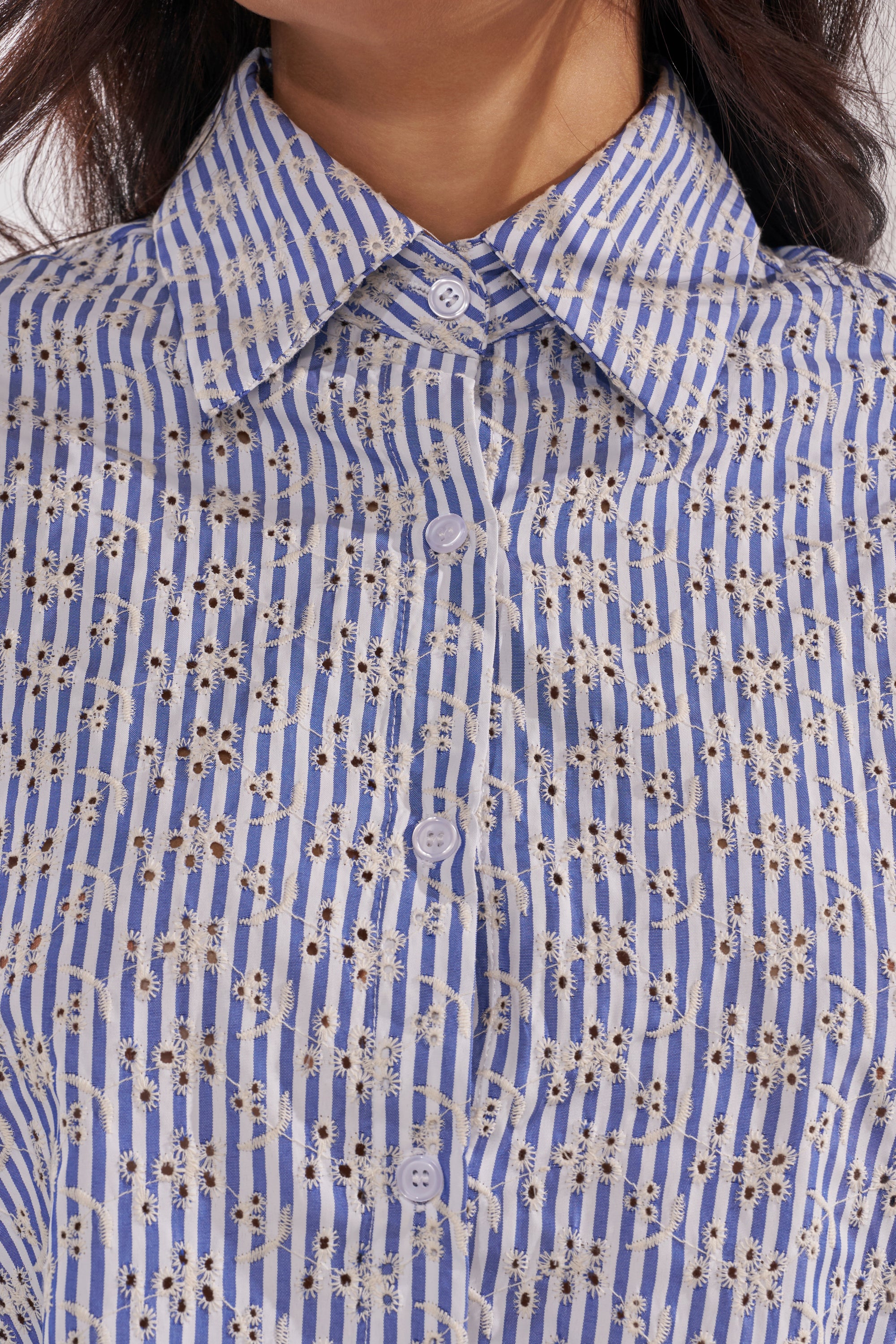 Close-up of a woman wearing the BLOSSOM CROP BUTTON DOWN, a blue and white striped shirt with delicate floral embroidery, pointed collar, and white front buttons. 8