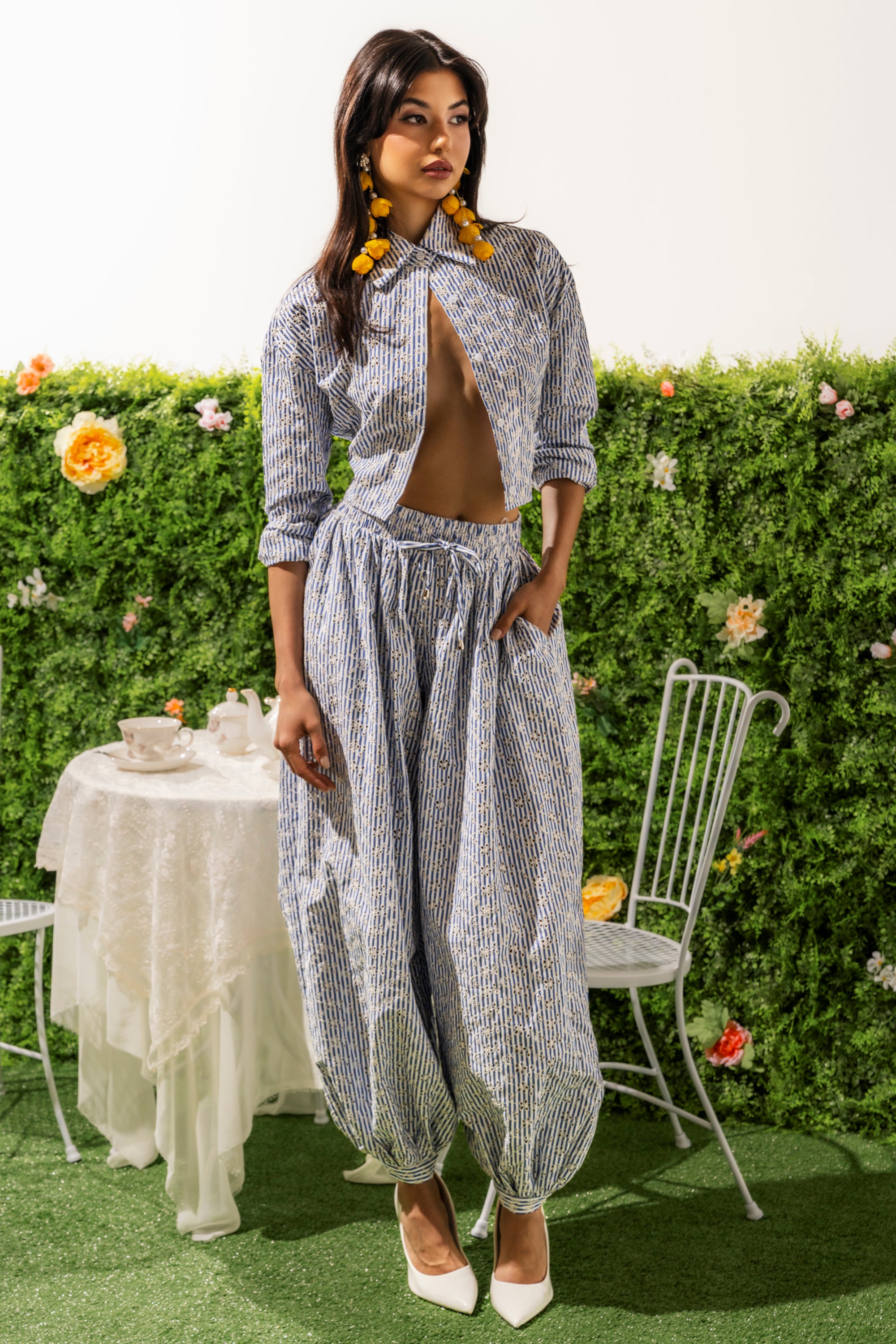 A woman poses by a green floral hedge, wearing the BLOSSOM BALLOON PANT as part of a blue and white striped set with a cropped top. She finishes the look with large yellow earrings, white heels, and stands near a table and chairs. 1