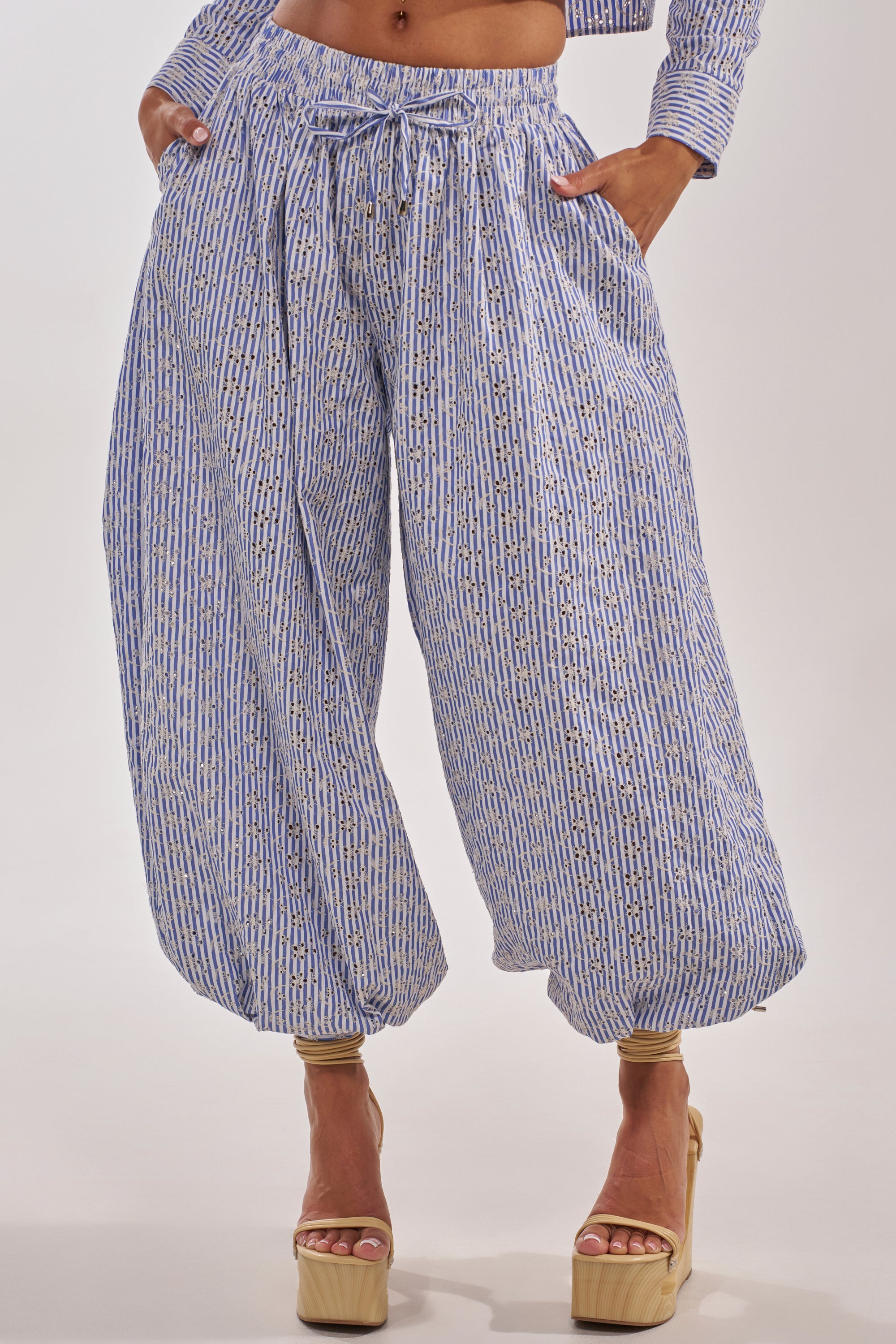 A person wears the BLOSSOM BALLOON PANT, featuring blue and white stripes, a wide-leg silhouette, drawstring waist, and pockets, styled with tan high platform heels. 0