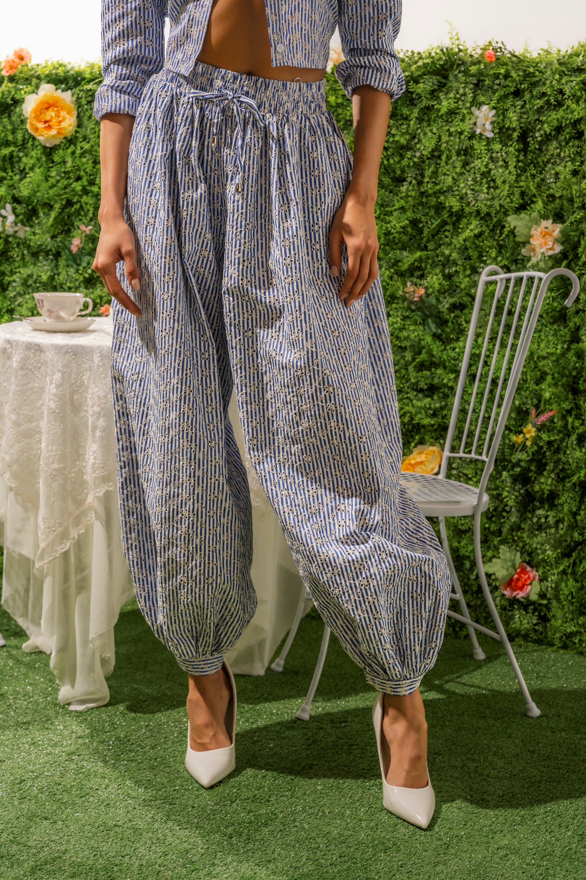 Wearing the BLOSSOM BALLOON PANT, featuring a blue-and-white pattern, a person in white heels stands on grass by a metal chair and table with a teacup, set before a hedge full of flowers. 2
