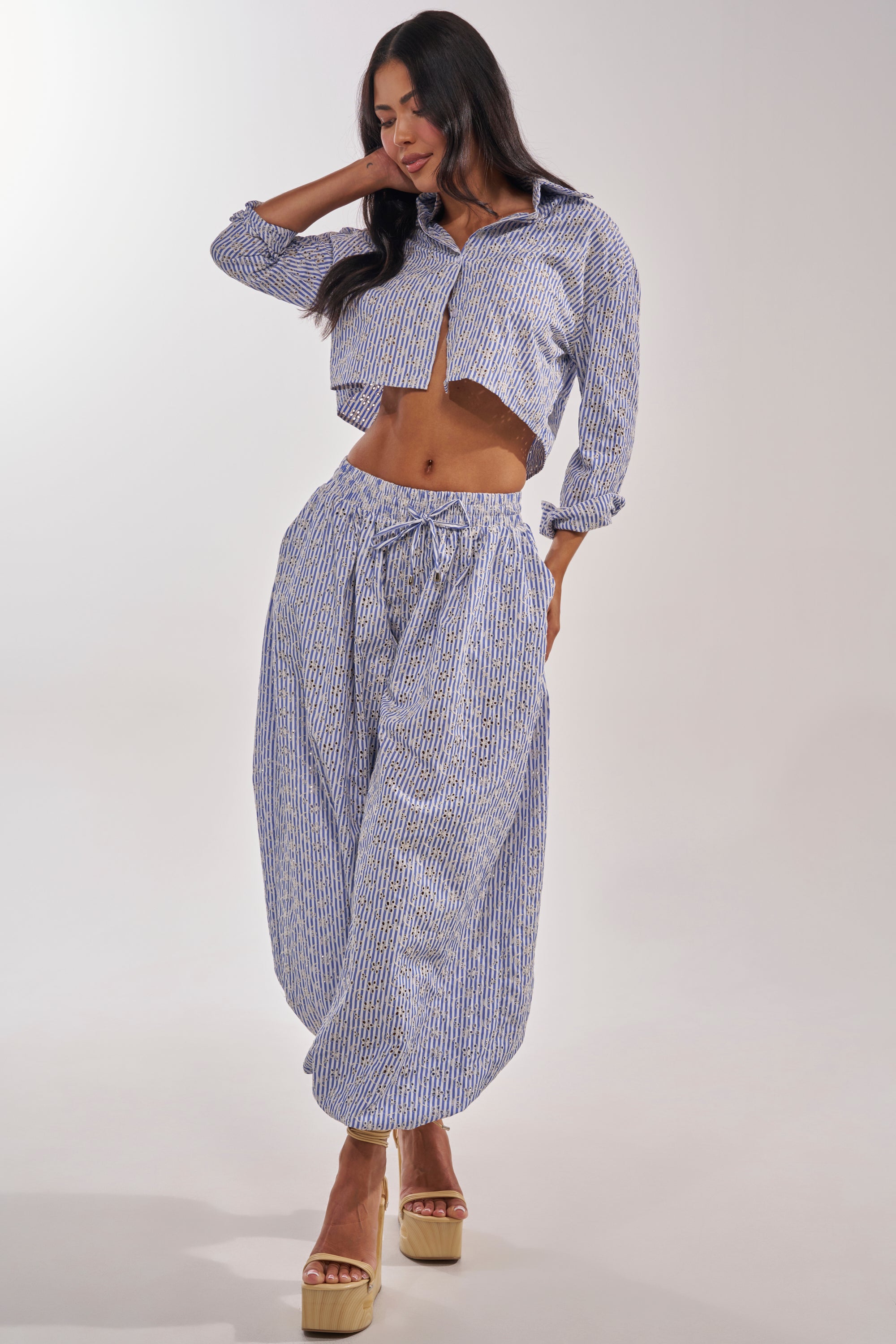 A woman models the BLOSSOM BALLOON PANT—a high-waisted, wide-leg blue and white striped pant—with a matching cropped button-up shirt and platform sandals. Her long dark hair flows against a plain, light background. 7