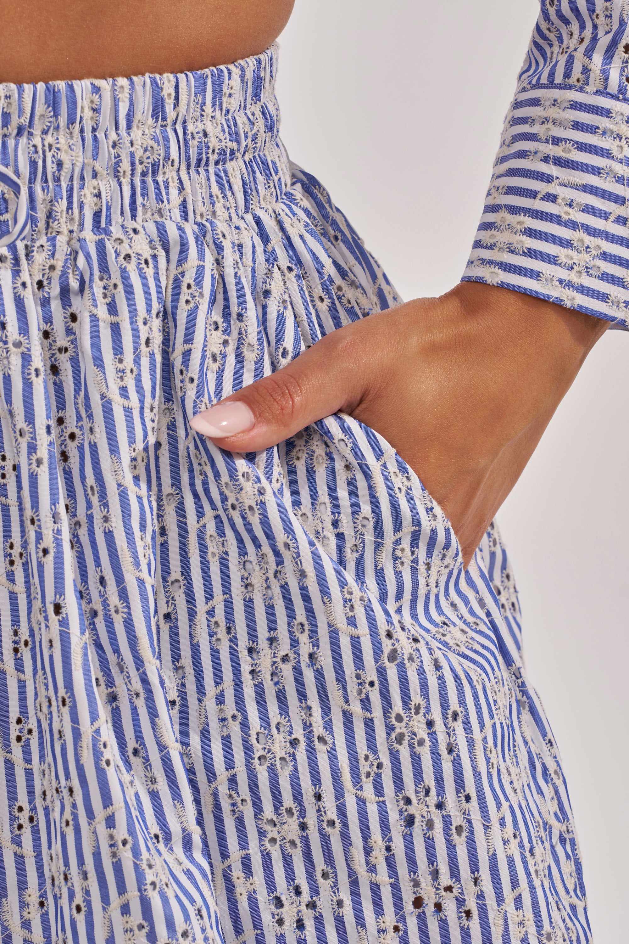 A close-up of a person’s hand in the pocket of the BLOSSOM BALLOON PANT, a blue and white vertically striped style with delicate floral embroidery, paired with a matching long-sleeve top. 8