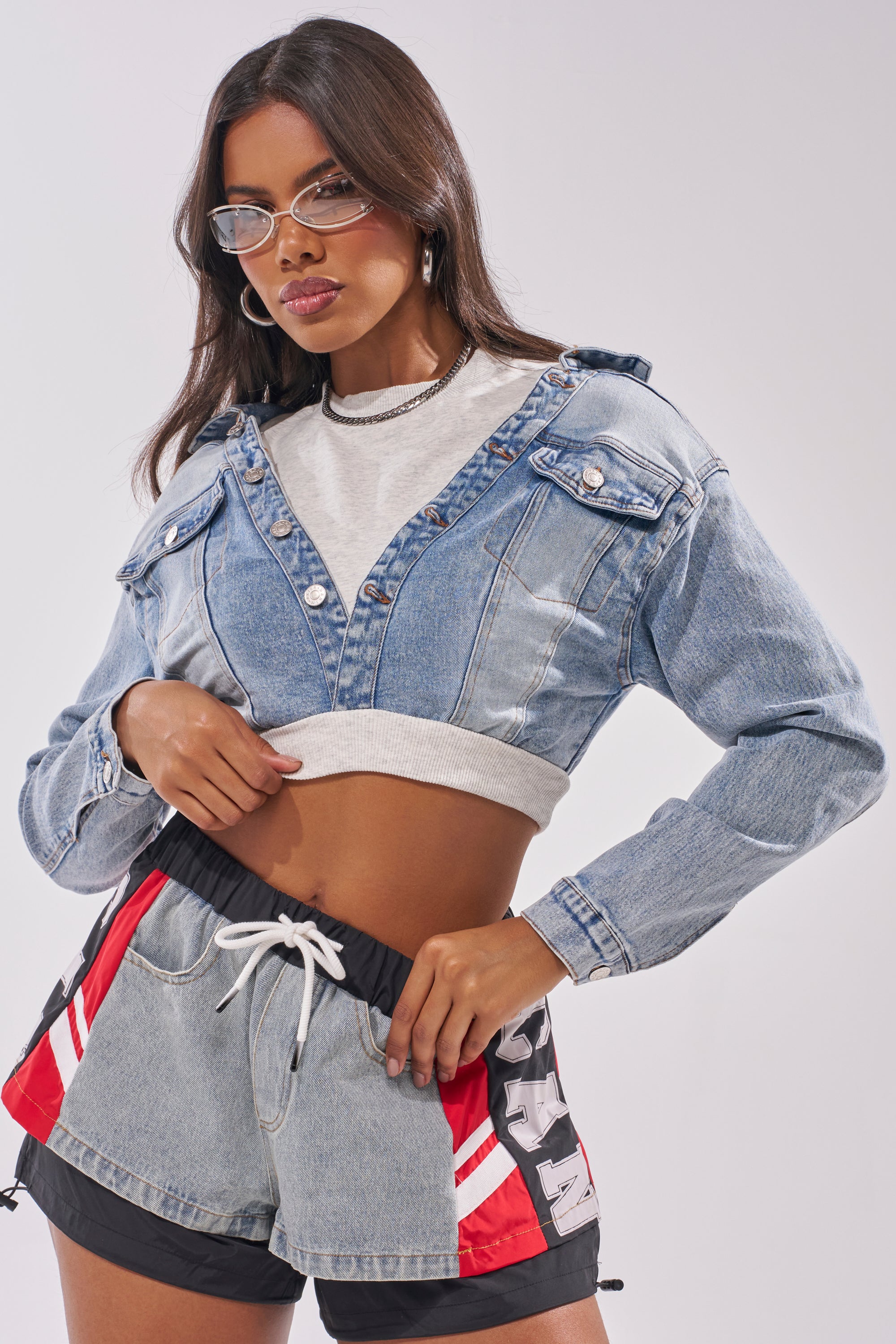 A woman confidently poses against a light background wearing clear glasses, the DOUBLE FUN SWEATSHIRT in gray, a cropped denim jacket, and denim athletic shorts featuring red, white, and black accents. 0