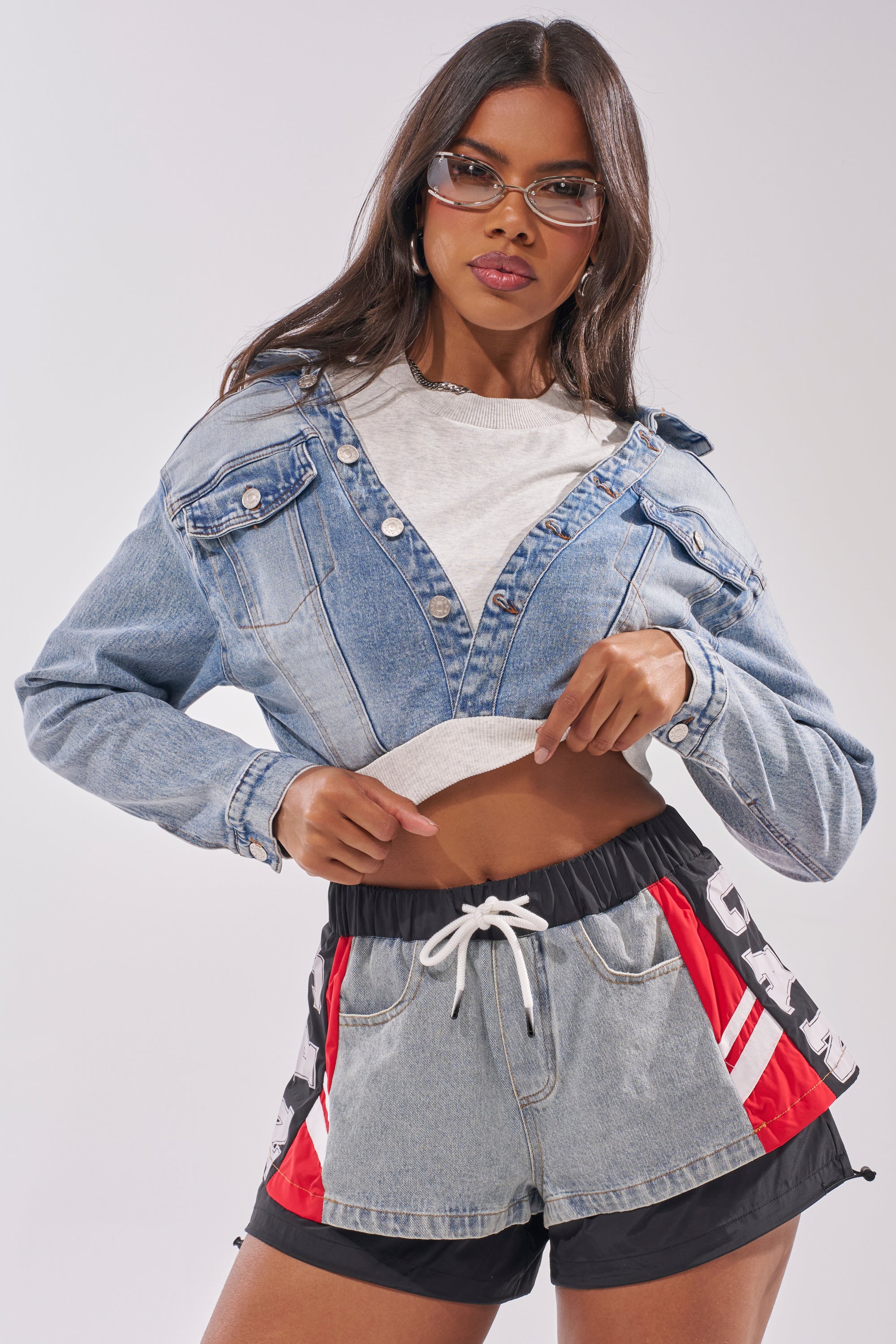 Woman wearing the DOUBLE FUN SWEATSHIRT layered under a mixed-media denim cropped jacket, styled with denim and black athletic shorts featuring red and white stripes. She accessorizes with glasses and wears her long hair loose. 1