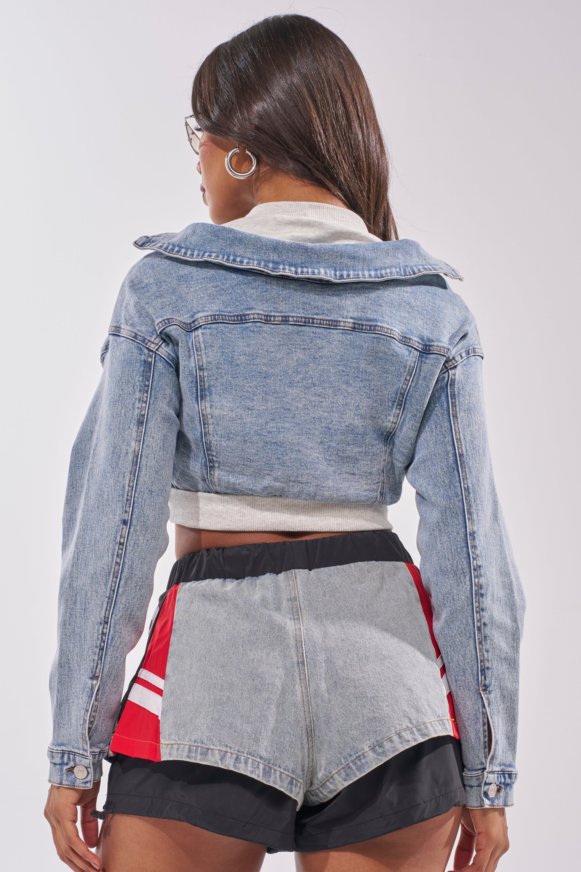 A woman with straight brown hair is seen from the back, wearing the DOUBLE FUN SWEATSHIRT, high-waisted denim, black athletic shorts with red and white stripes, and hoop earrings. 3