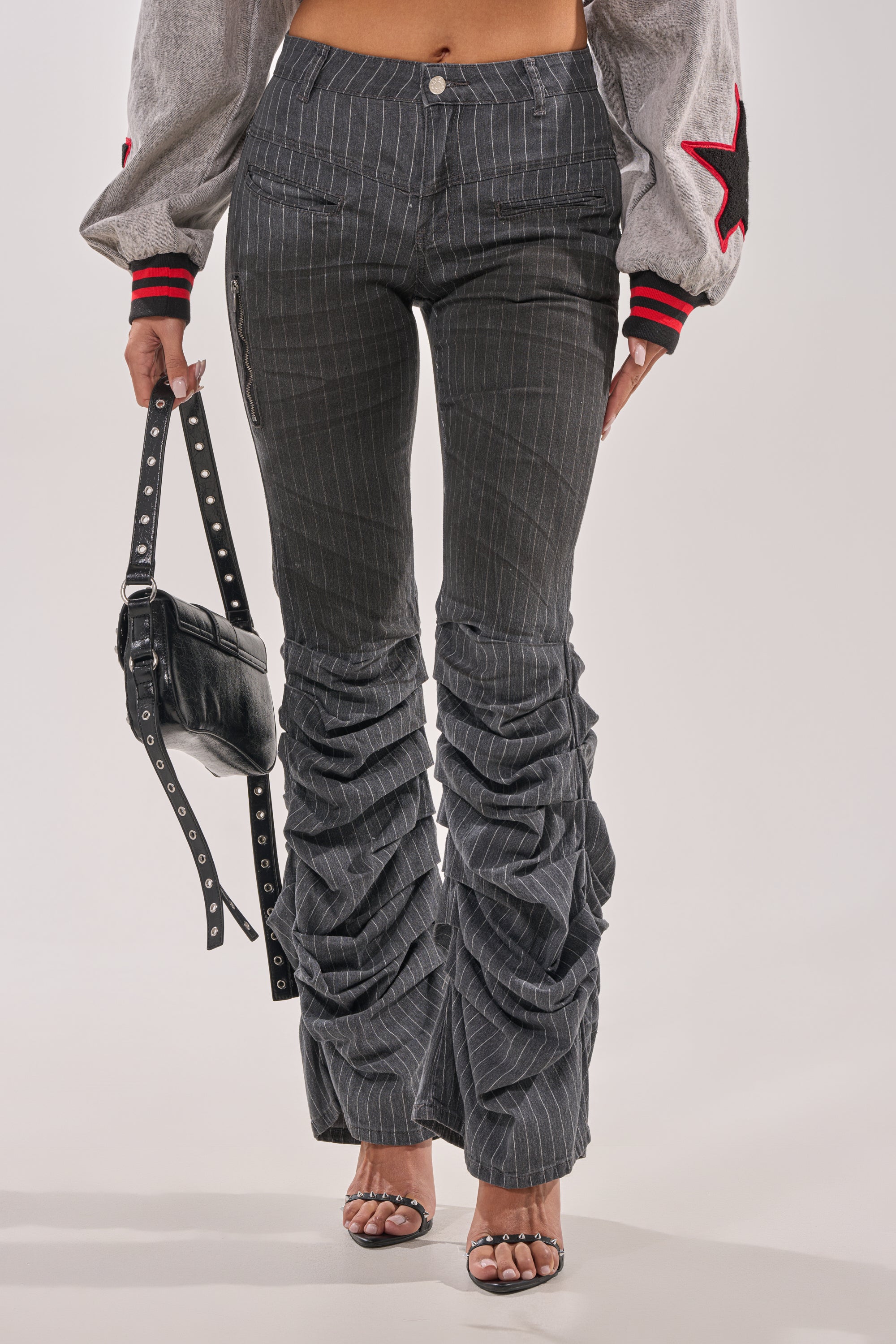 Wearing the TALK TO ME STRIPE TROUSER, a cropped jacket with red and black star sleeve details, a studded black handbag, and open-toe heeled sandals. 0