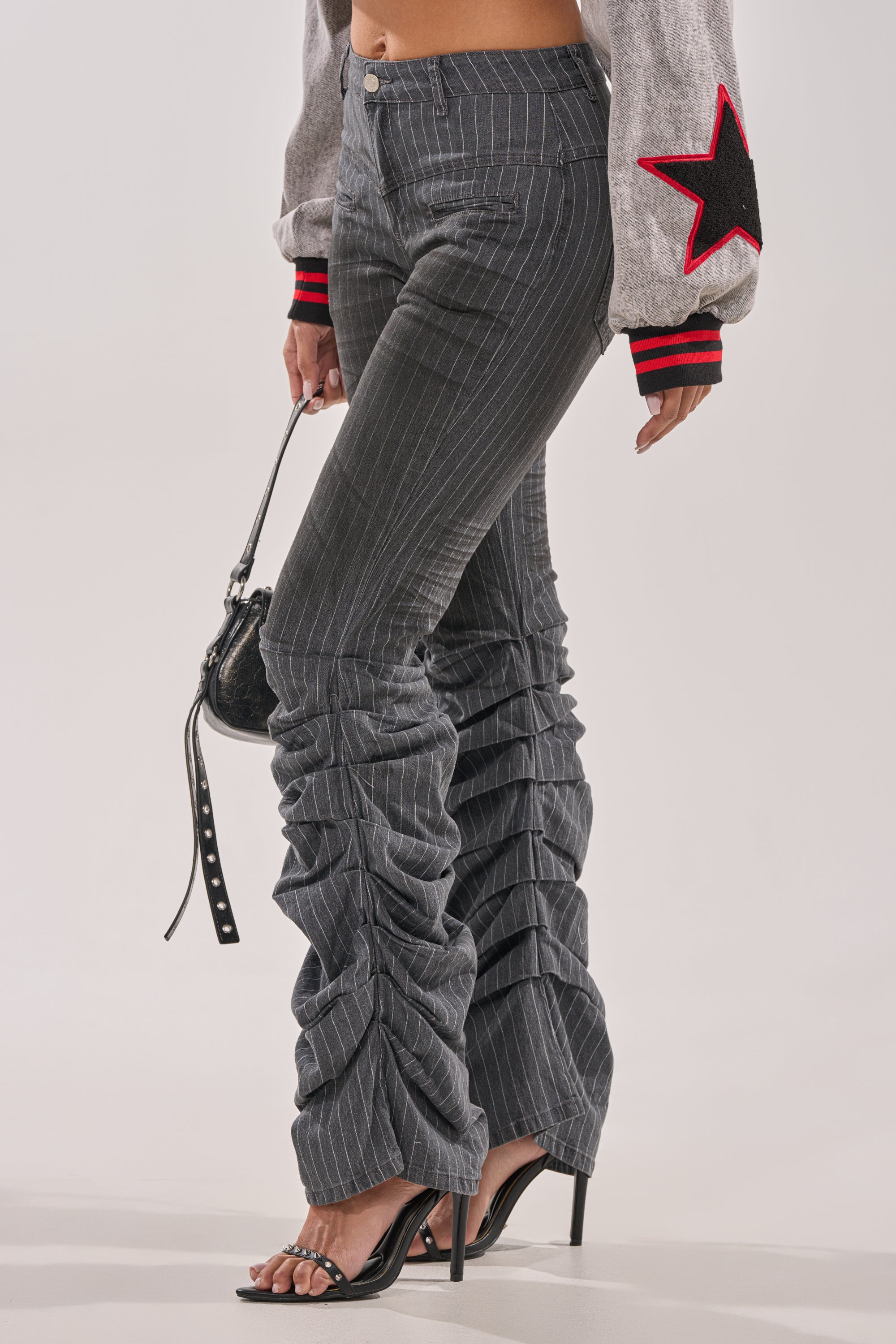 A person wearing TALK TO ME STRIPE TROUSER pinstripe flare pants with a high-rise waist, black high-heeled sandals, a gray sweater with red and black star sleeve details, and a small black handbag. 1