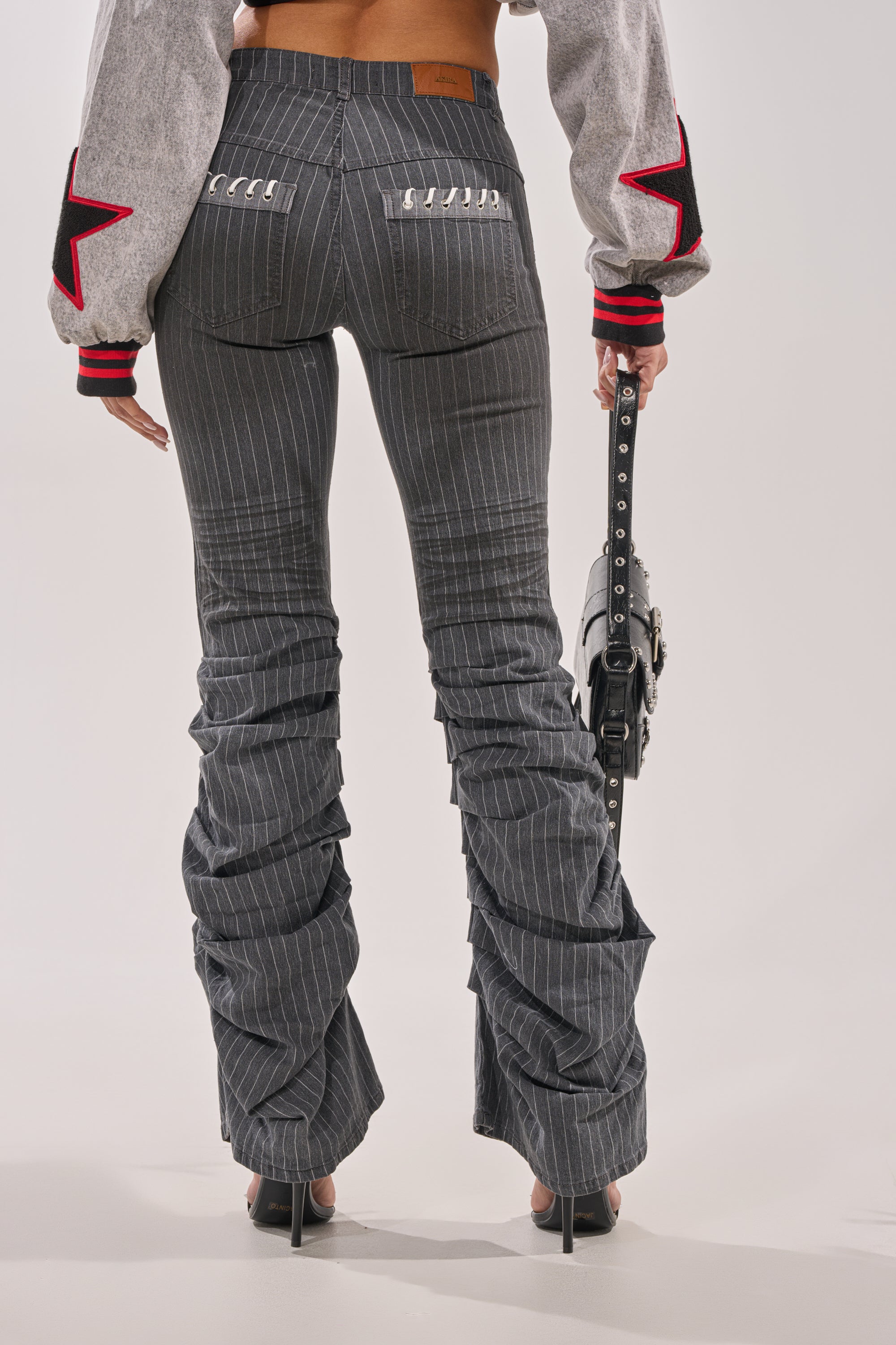 A person seen from behind wears the TALK TO ME STRIPE TROUSER—gray pinstripe ruched flare pants with a high-rise waist—paired with a red-star cropped jacket, black handbag, and black high heels. 2