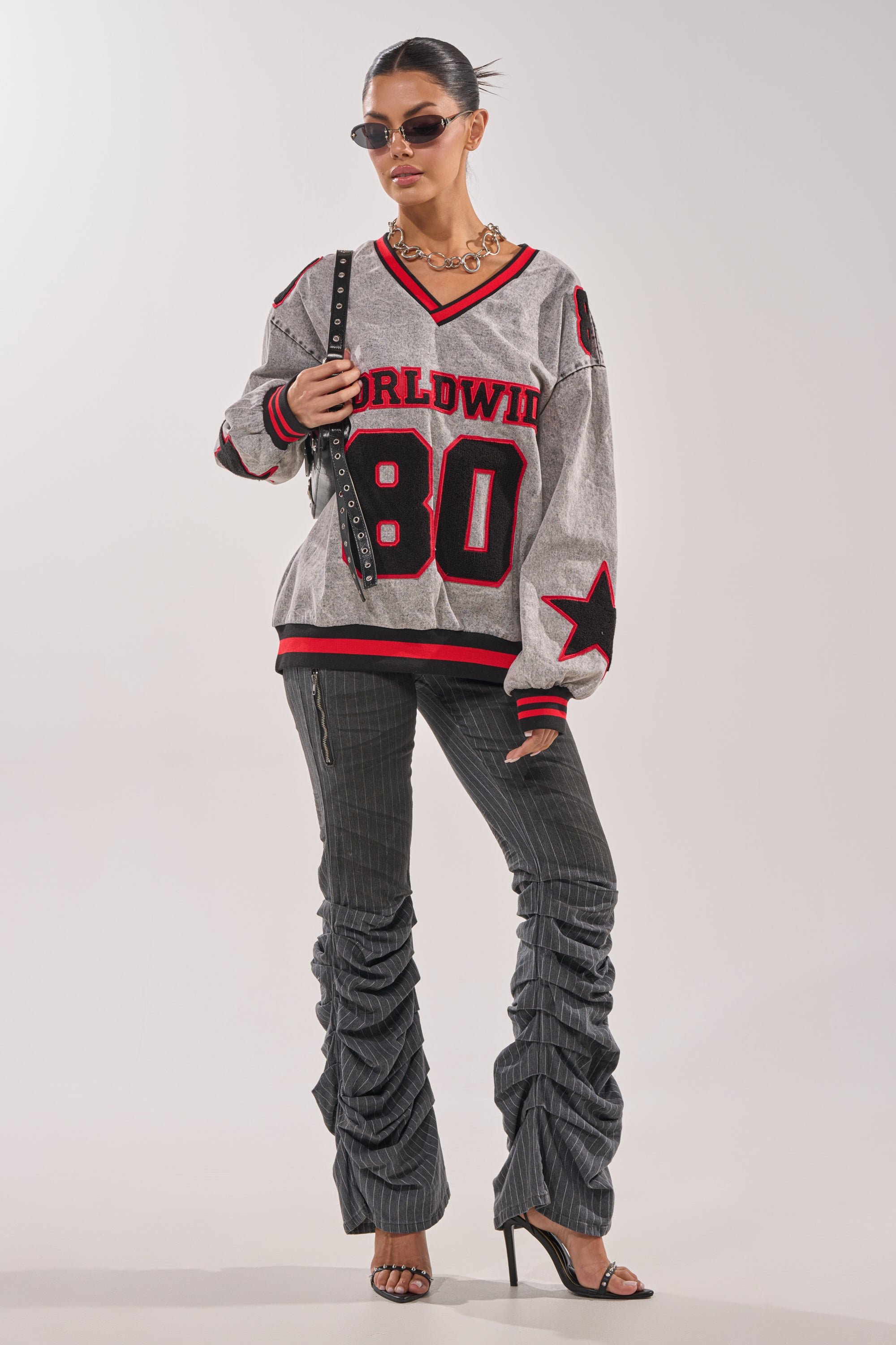 A woman poses confidently in sunglasses, a chunky necklace, a loose grey and red "Worldwind 80" jersey, TALK TO ME STRIPE TROUSER pinstripe flare pants, black heels, and a small studded purse against a plain background. 5