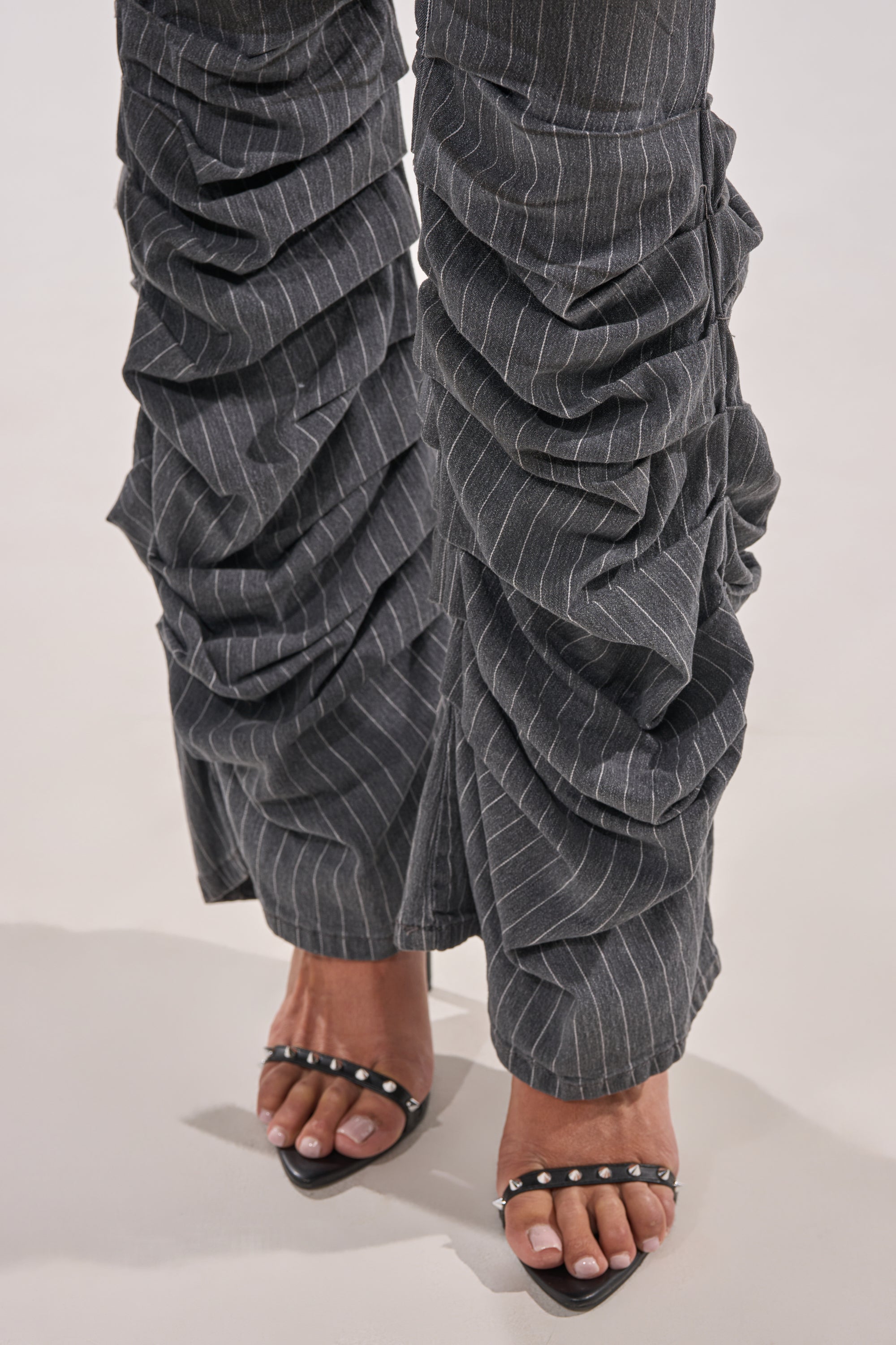 A close-up of a person wearing TALK TO ME STRIPE TROUSER—grey pinstripe flare pants with ruched details and a high-rise waist—paired with black open-toe heels with silver studs, standing on a light background. 6