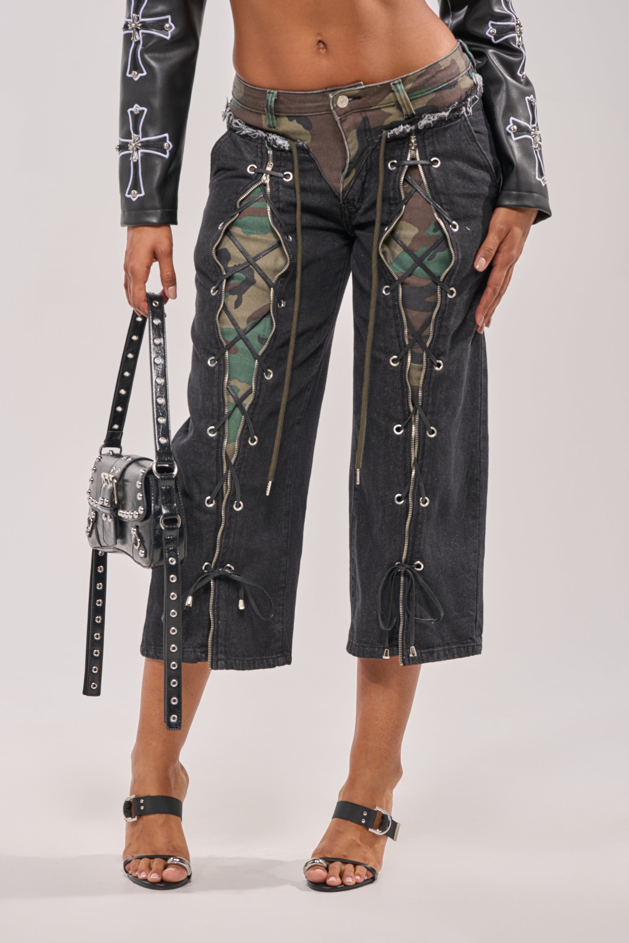 A person wears WALK THAT WALK CAPRI PANT featuring lace-up accents and camo mesh panels, styled with a studded mini bag, black leather jacket with white crosses, and black heeled sandals.