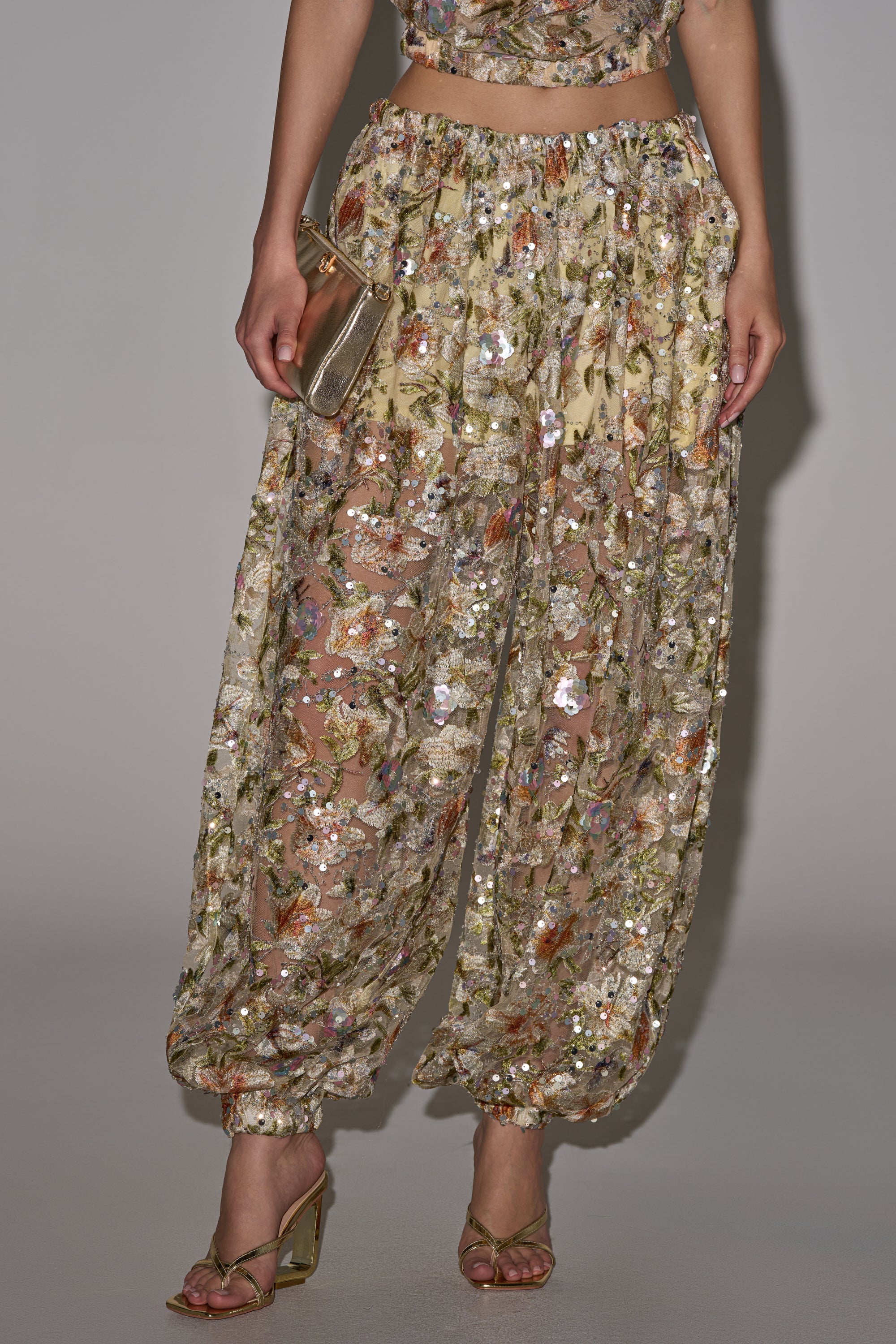 Wearing the LETS TALK ABOUT IT BALLOON PANT—sheer, ankle-gathered with floral embroidery and sequins—paired with gold heels and a metallic clutch, partially revealing the legs; upper body is partly visible. 0