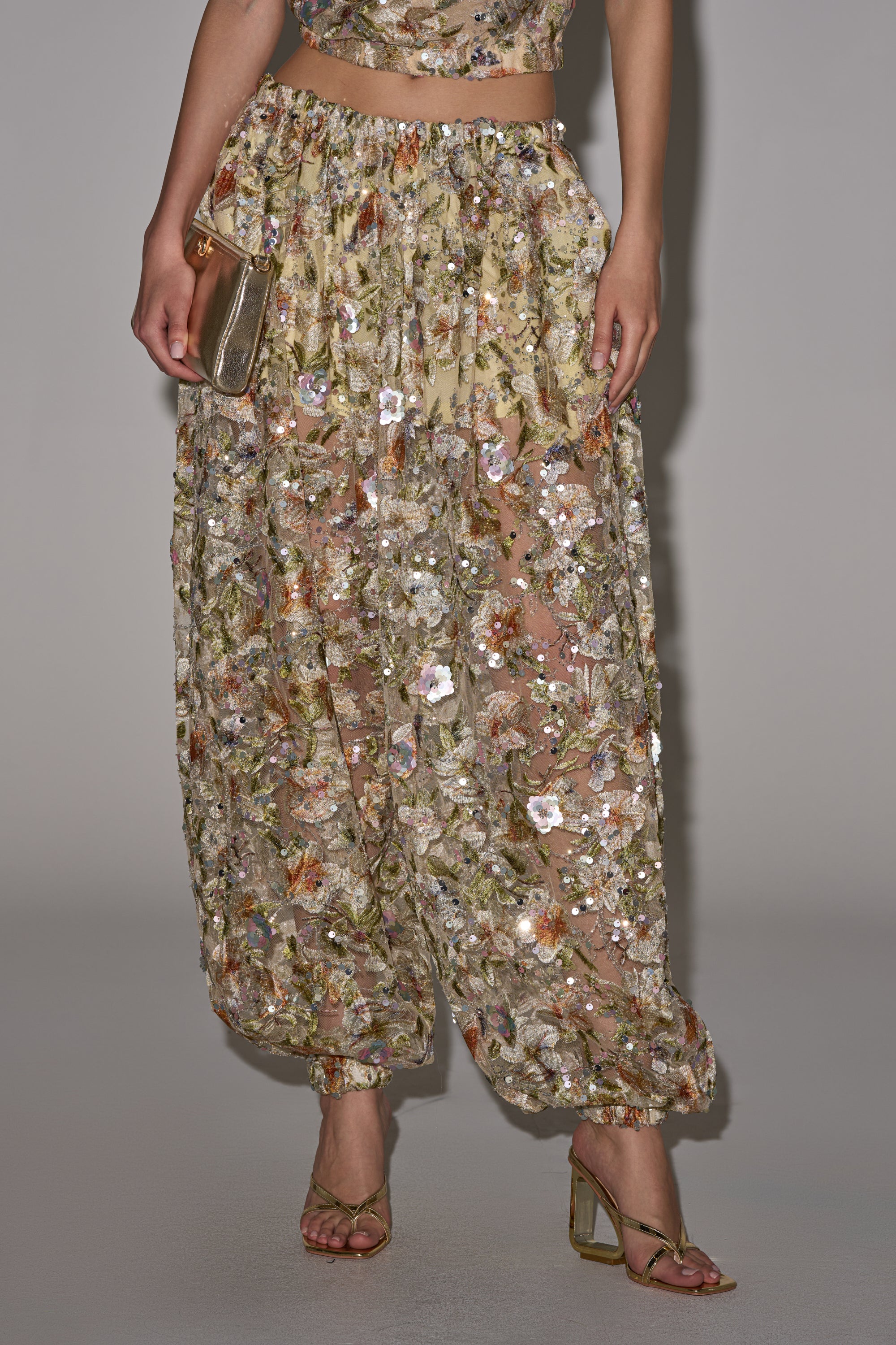 A person wears the LETS TALK ABOUT IT BALLOON PANT, featuring sheer fabric with floral embroidery and sequins, paired with gold high-heeled sandals and a small metallic clutch against a plain background. 2