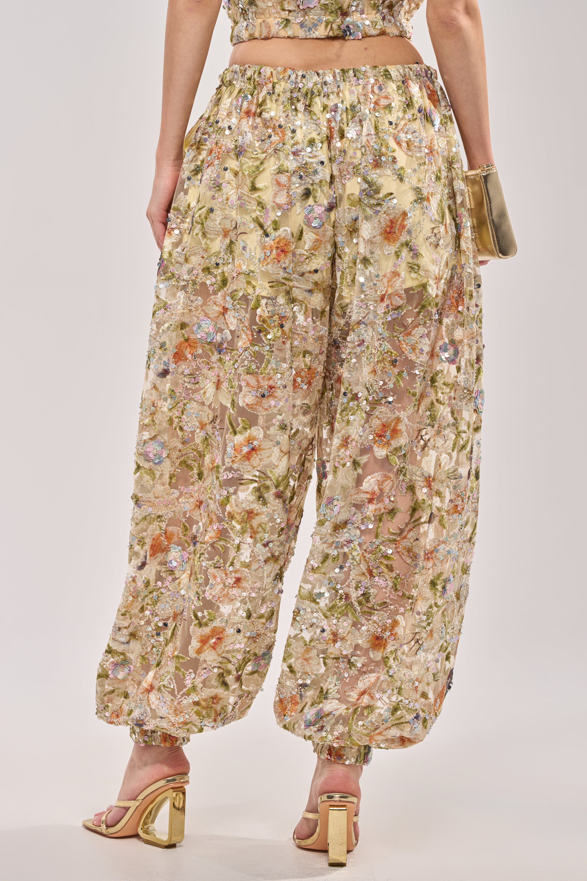 Photographed from the back, a person wears the LETS TALK ABOUT IT BALLOON PANT—loose, sheer pants with an elastic waistband and cuffs—styled with gold high-heeled sandals and a matching gold clutch. 4