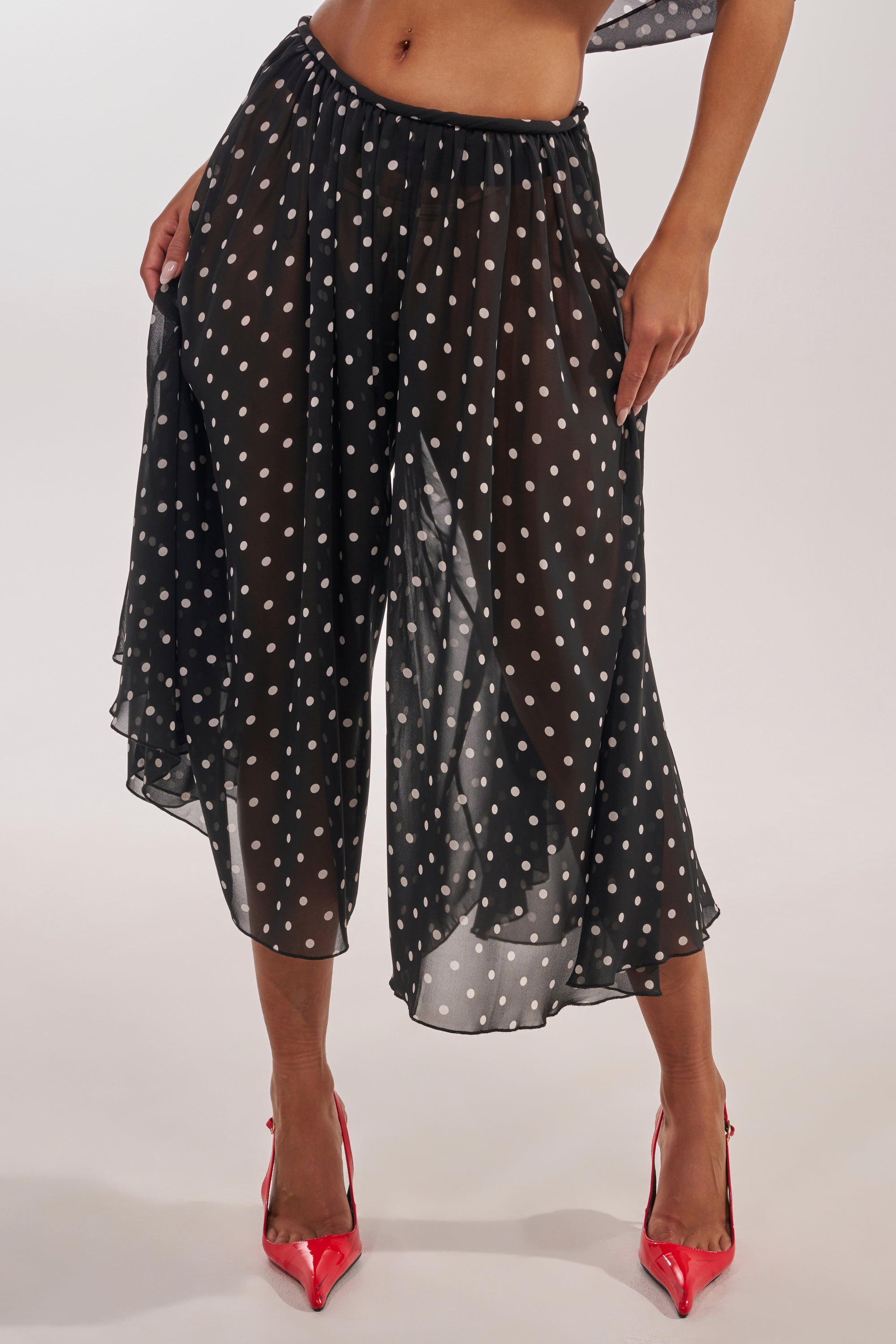 Model wears the COOL GIRL POLKA DOT GAUCHO PANT in sheer black, paired with bright red pointed heels, standing with hands on hips against a plain background. 0