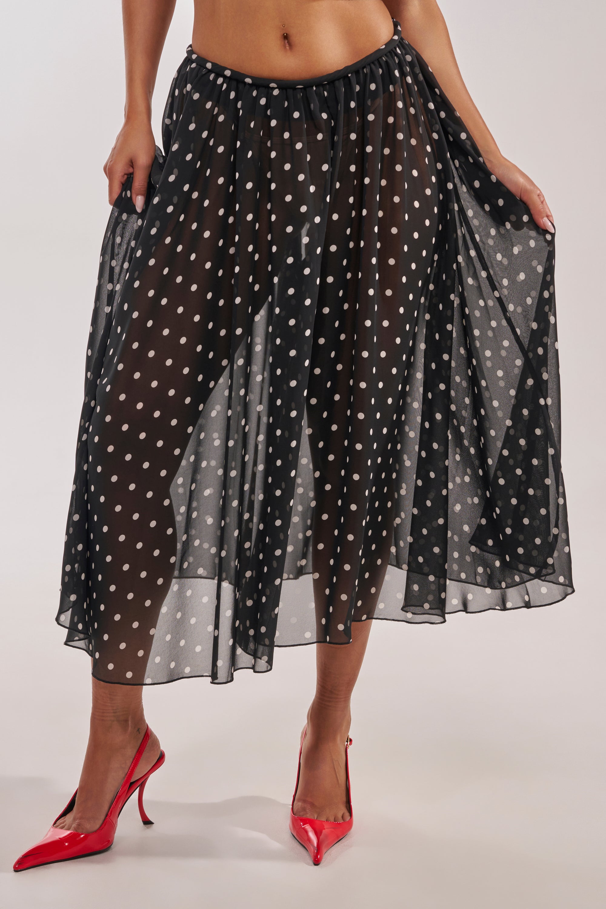 A person wears the COOL GIRL POLKA DOT GAUCHO PANT, styled with bright red pointed heels, standing on a light background and holding out the sides to showcase the flowing polka dot design. 1