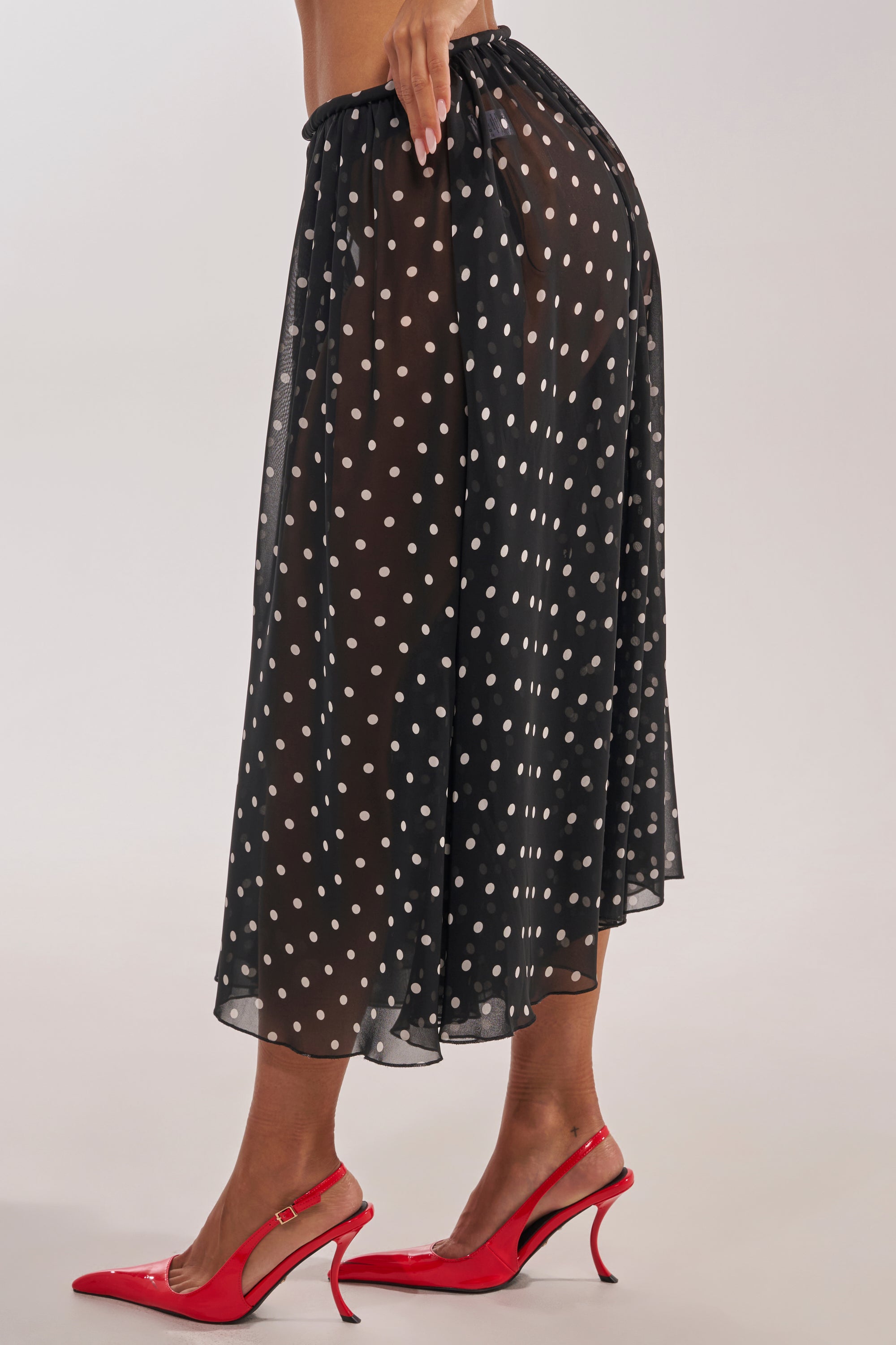 A person models the COOL GIRL POLKA DOT GAUCHO PANT in sheer black with polka dots and bright red heels, shown from the side to highlight the flowy fabric, chic style, and the wearer’s legs. 2