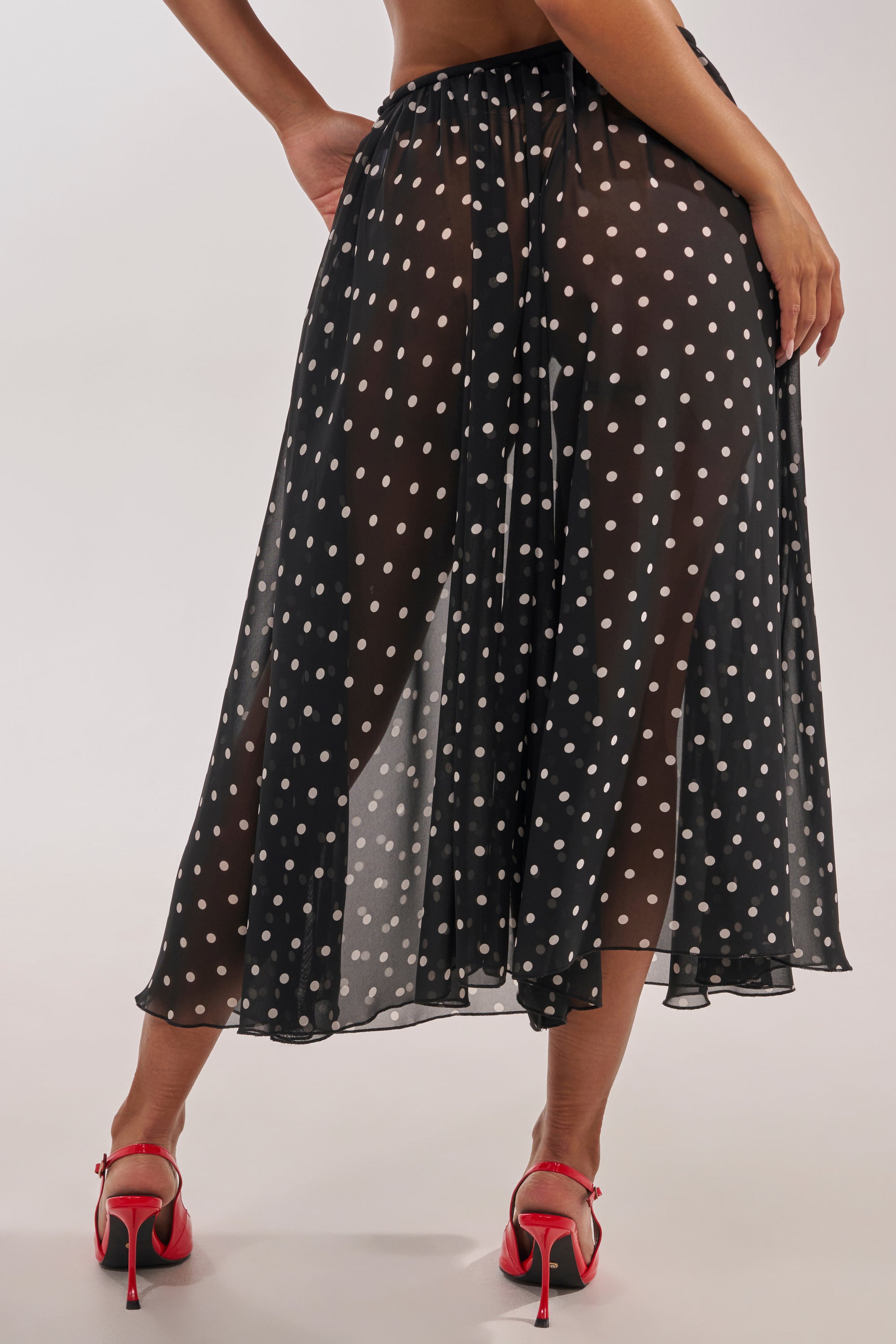A woman, seen from behind against a neutral background, wears red heels and the COOL GIRL POLKA DOT GAUCHO PANT—a sheer black midi gaucho style with white polka dots that highlights her legs underneath. 3
