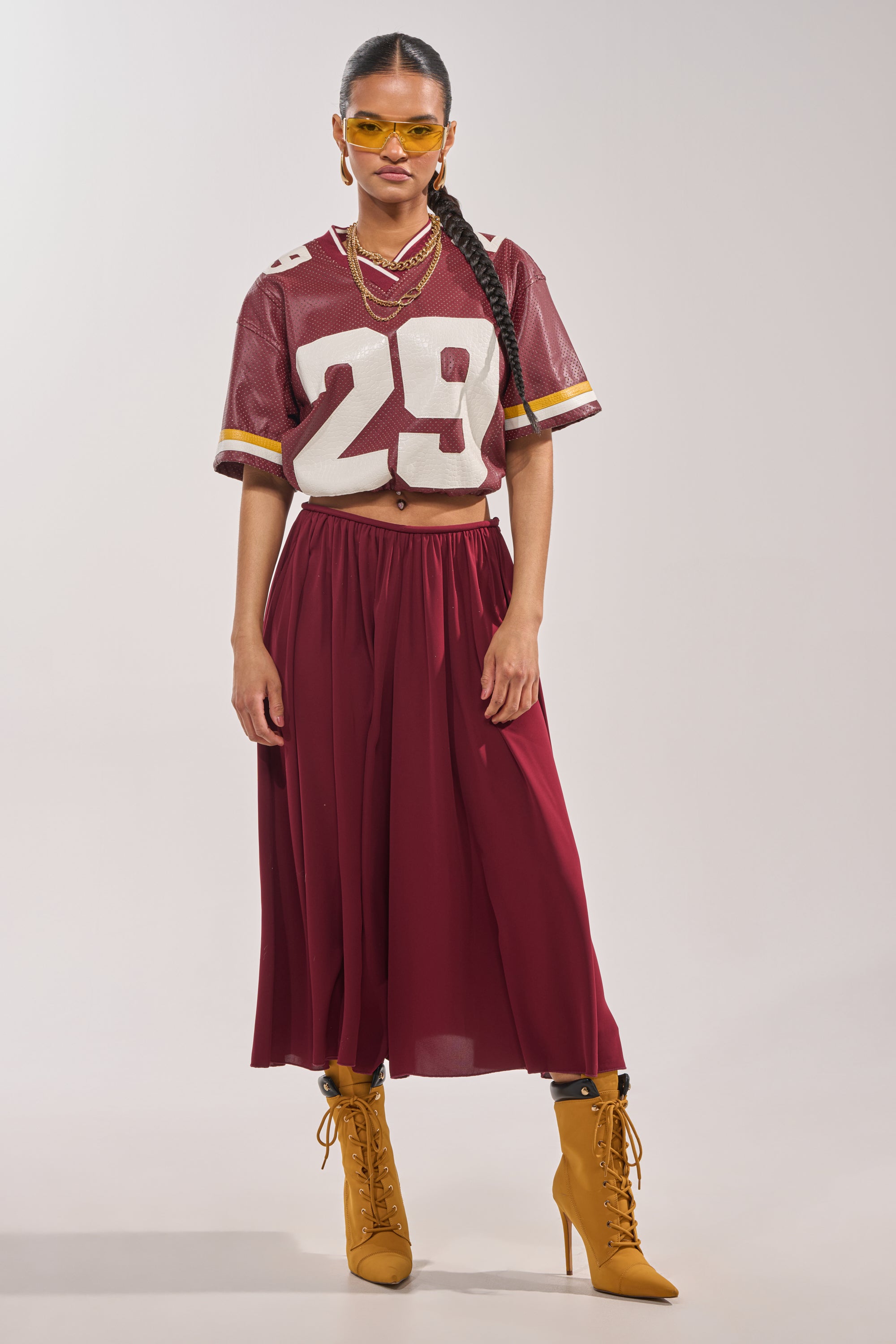 A woman wearing yellow sunglasses, a wine-hued football jersey with the number 29, COOL GIRL GAUCHO PANT, and yellow lace-up boots stands against a plain background. 0