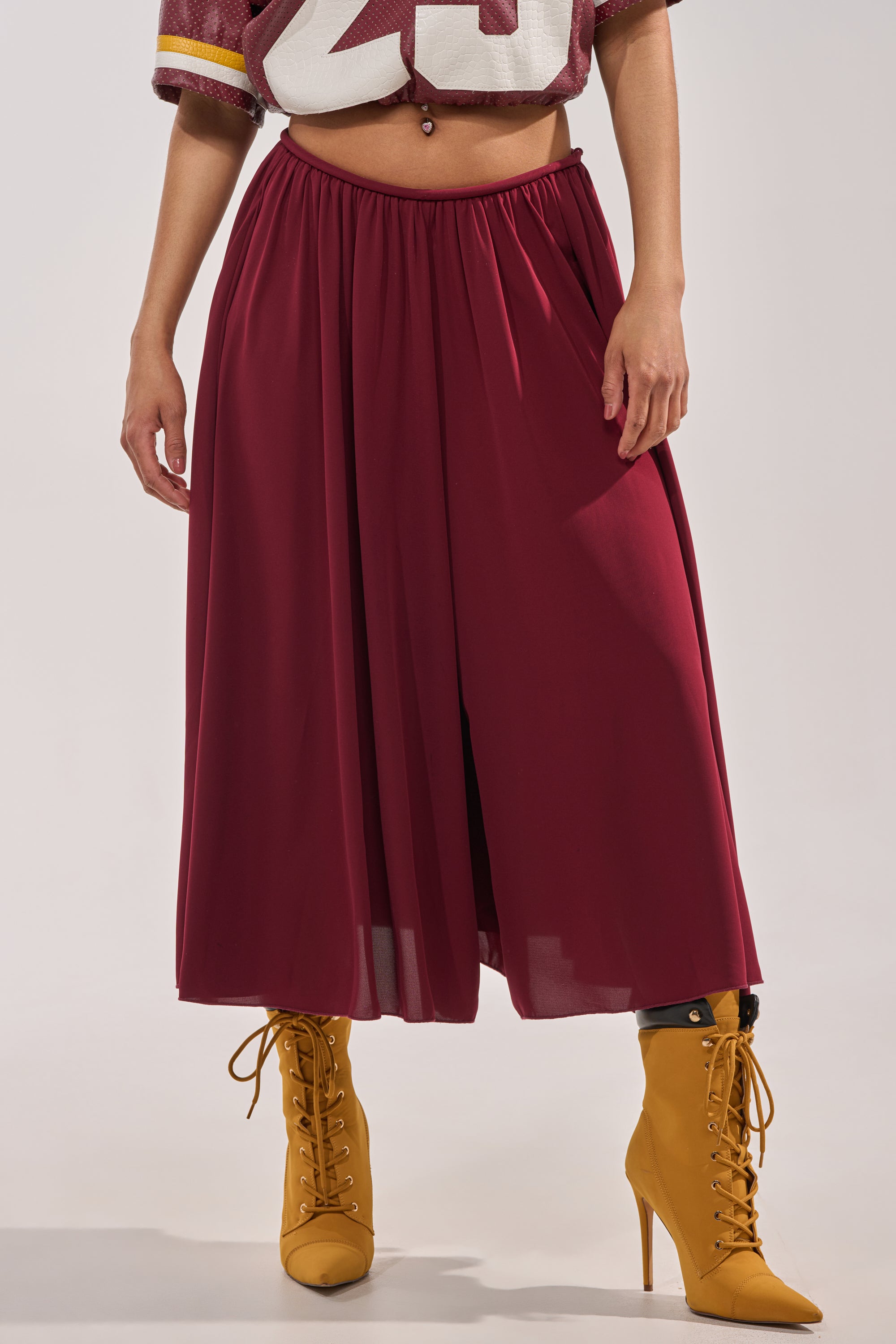A person wearing the COOL GIRL GAUCHO PANT in a wine hue with a high-rise waistband, paired with a cropped sports jersey and mustard yellow lace-up heeled boots. Photo is cropped from shoulders down. 1