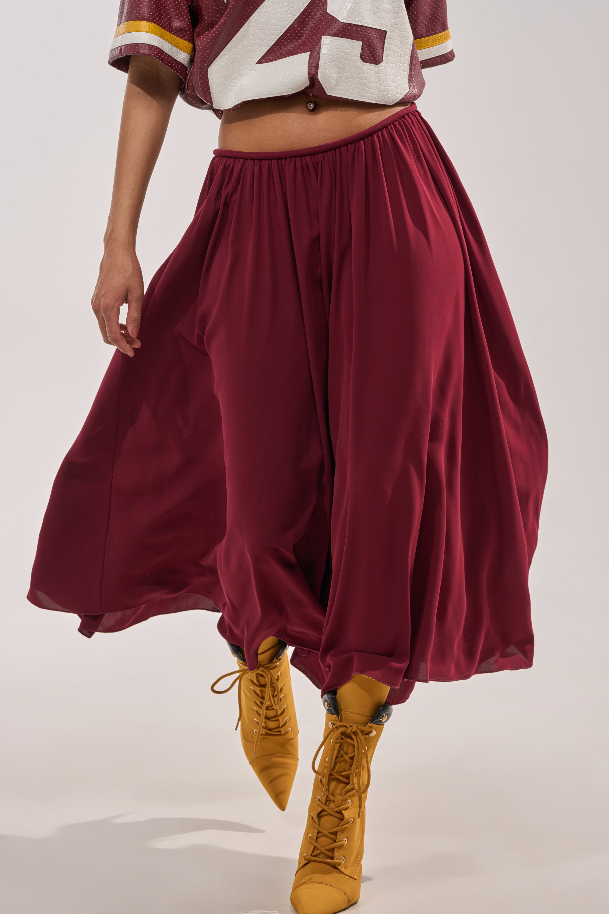 A person in wine-hued COOL GIRL GAUCHO PANT, a burgundy sports jersey, and mustard yellow lace-up boots walks forward. The image is cropped at the shoulders. 2