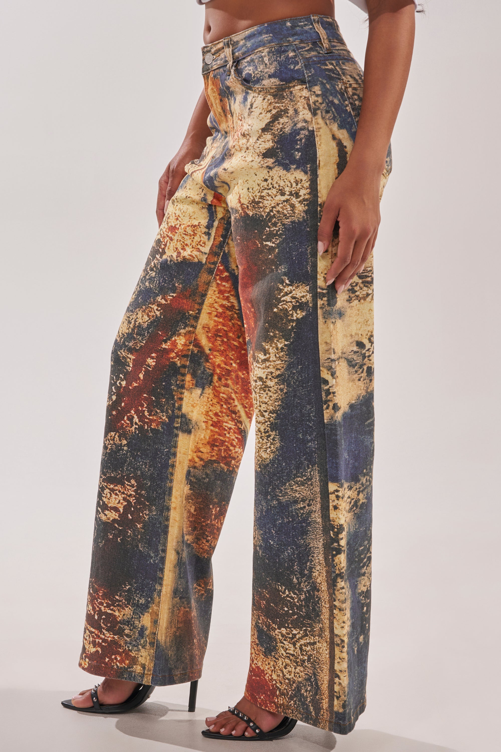 A person wears the POWER SQUAD RELAXED DENIM—wide-leg jeans featuring a bold abstract design in yellow, orange, blue, and black—paired with black open-toe heels. The image is cropped from waist to feet. 1