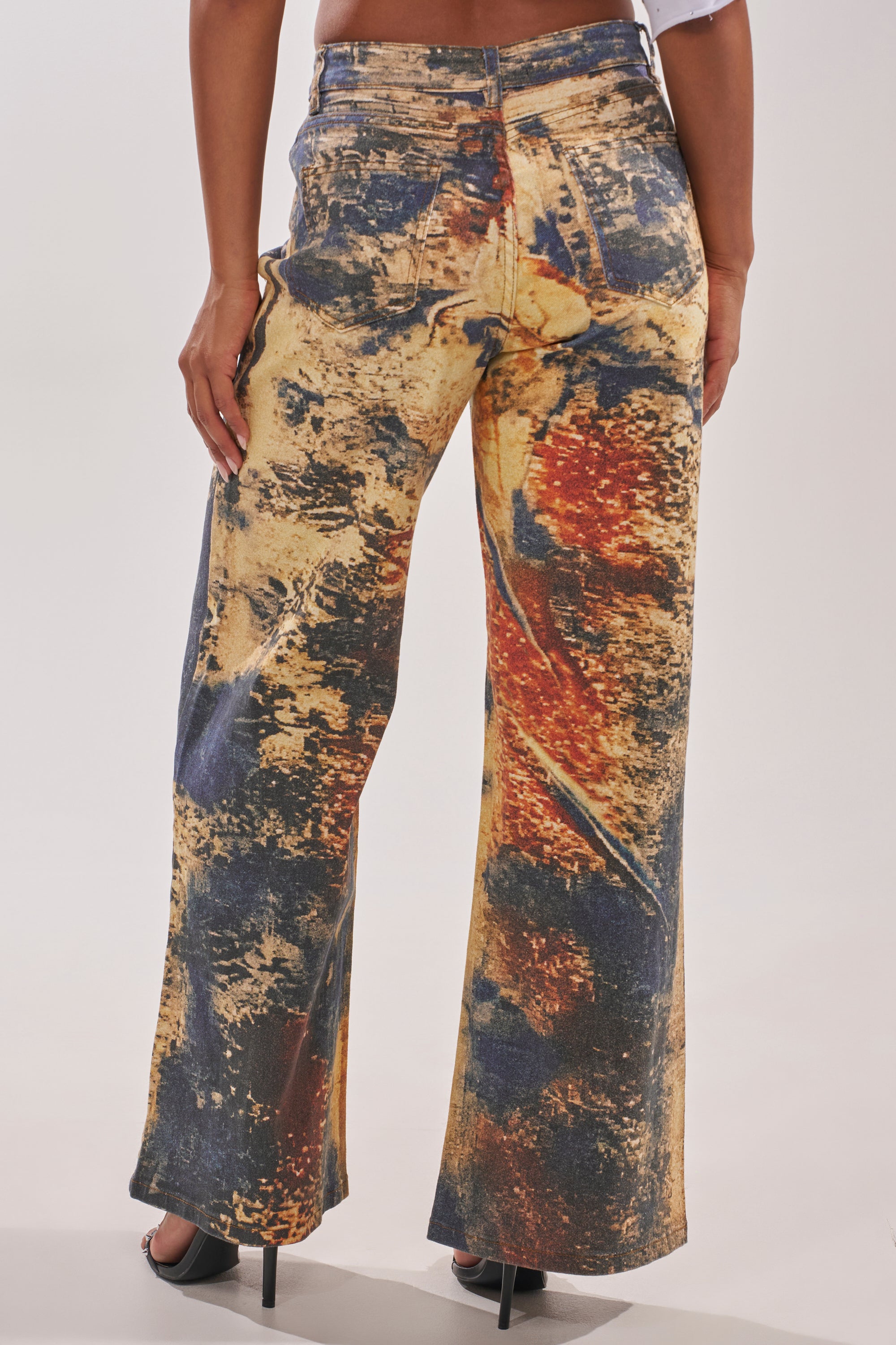 A person stands with their back to the camera, wearing POWER SQUAD RELAXED DENIM—high-rise, wide-leg jeans featuring a bold multicolored abstract print in yellow, orange, blue, and black—stylishly paired with black heeled shoes. 2