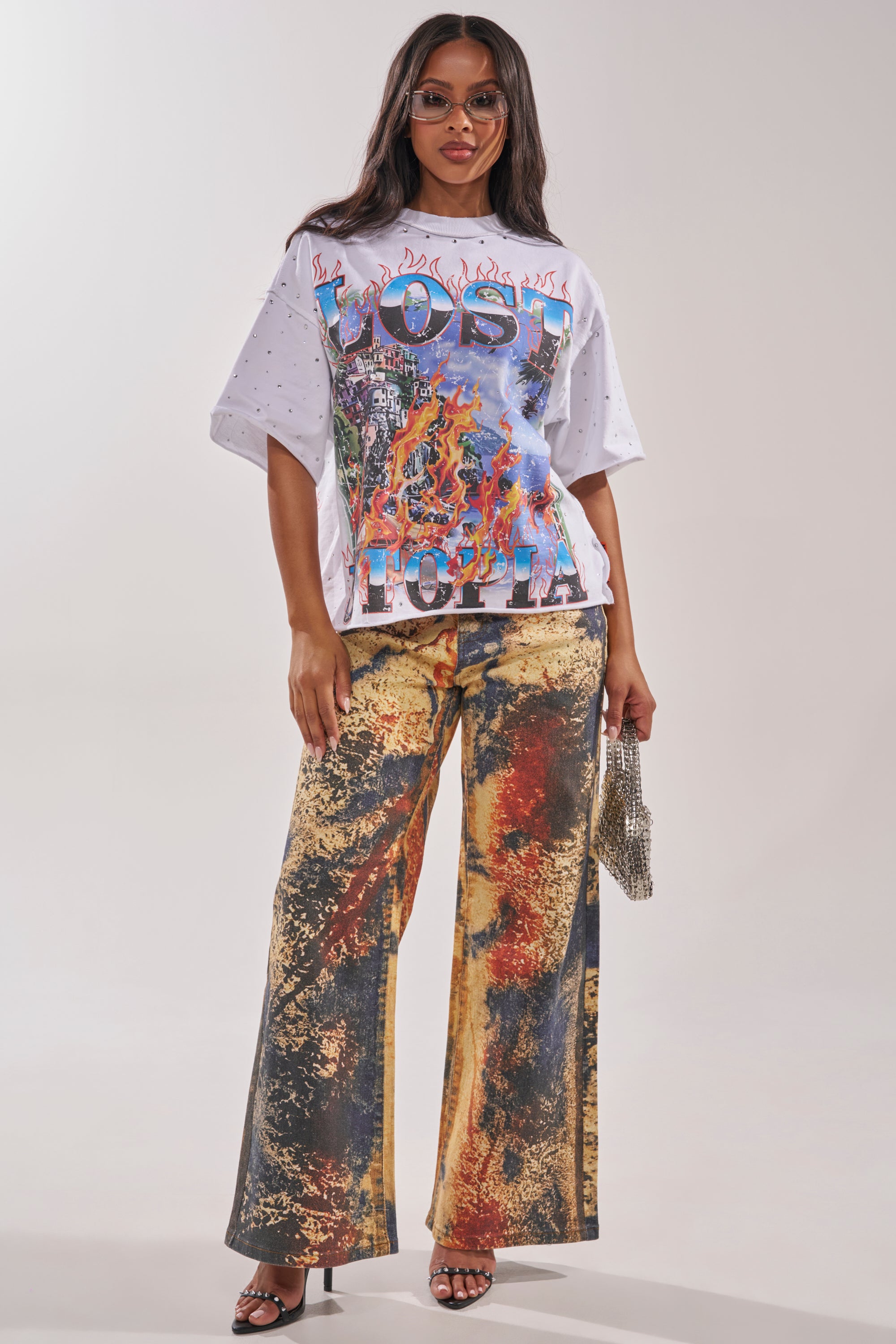 A woman wears sunglasses, a "LOST" graphic tee, POWER SQUAD RELAXED DENIM jeans with an abstract wide-leg style, black heeled sandals, and carries a small silver purse. 3