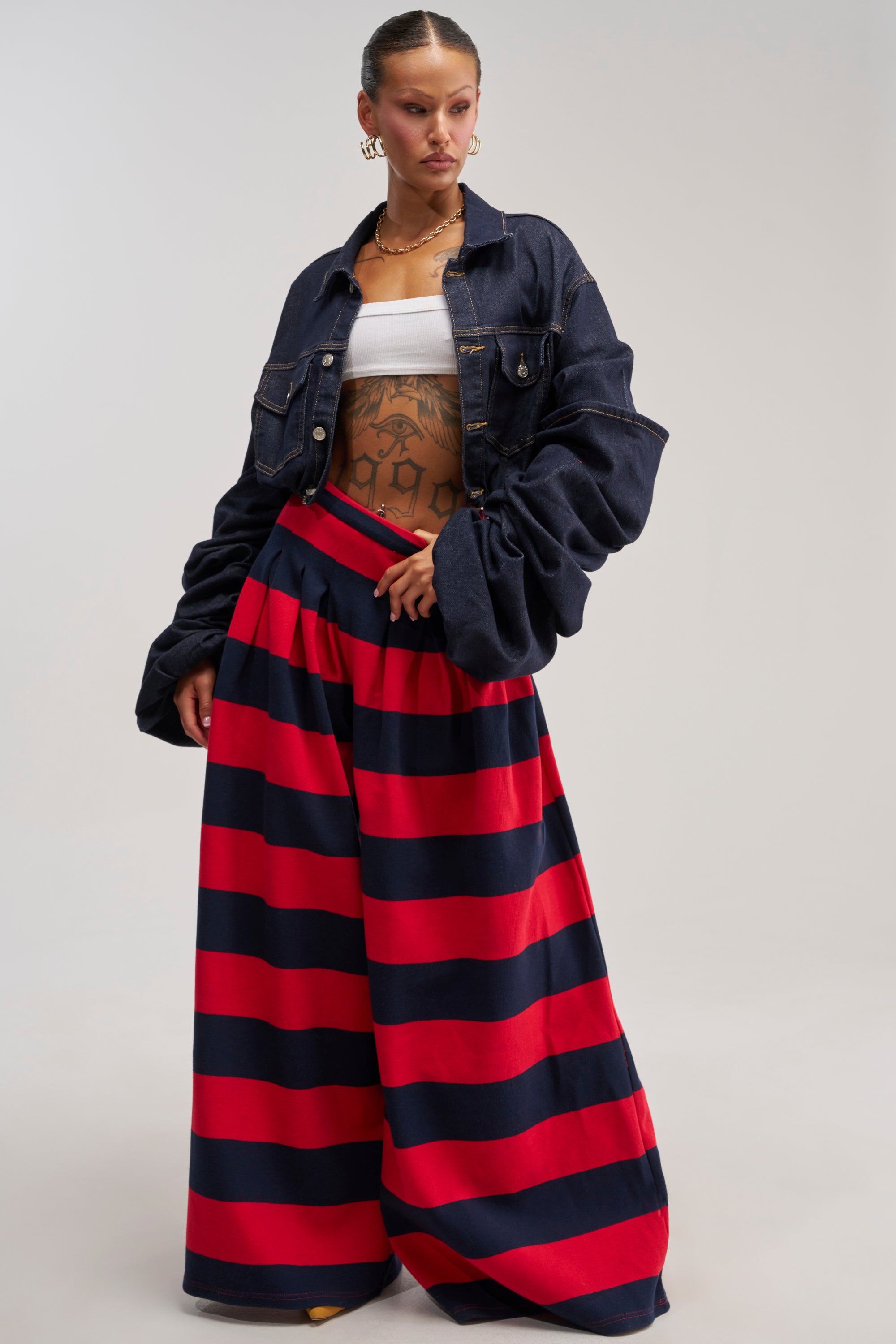 A woman stands confidently in the SILVER COAST RED STRIPE TROUSER, featuring a wide-leg silhouette with bold red and navy stripes. She pairs it with a dark denim jacket and a white tube top, accessorizing her look with hoop earrings and displaying tattoos on her stomach. Her hair is slicked back for a sleek finish.