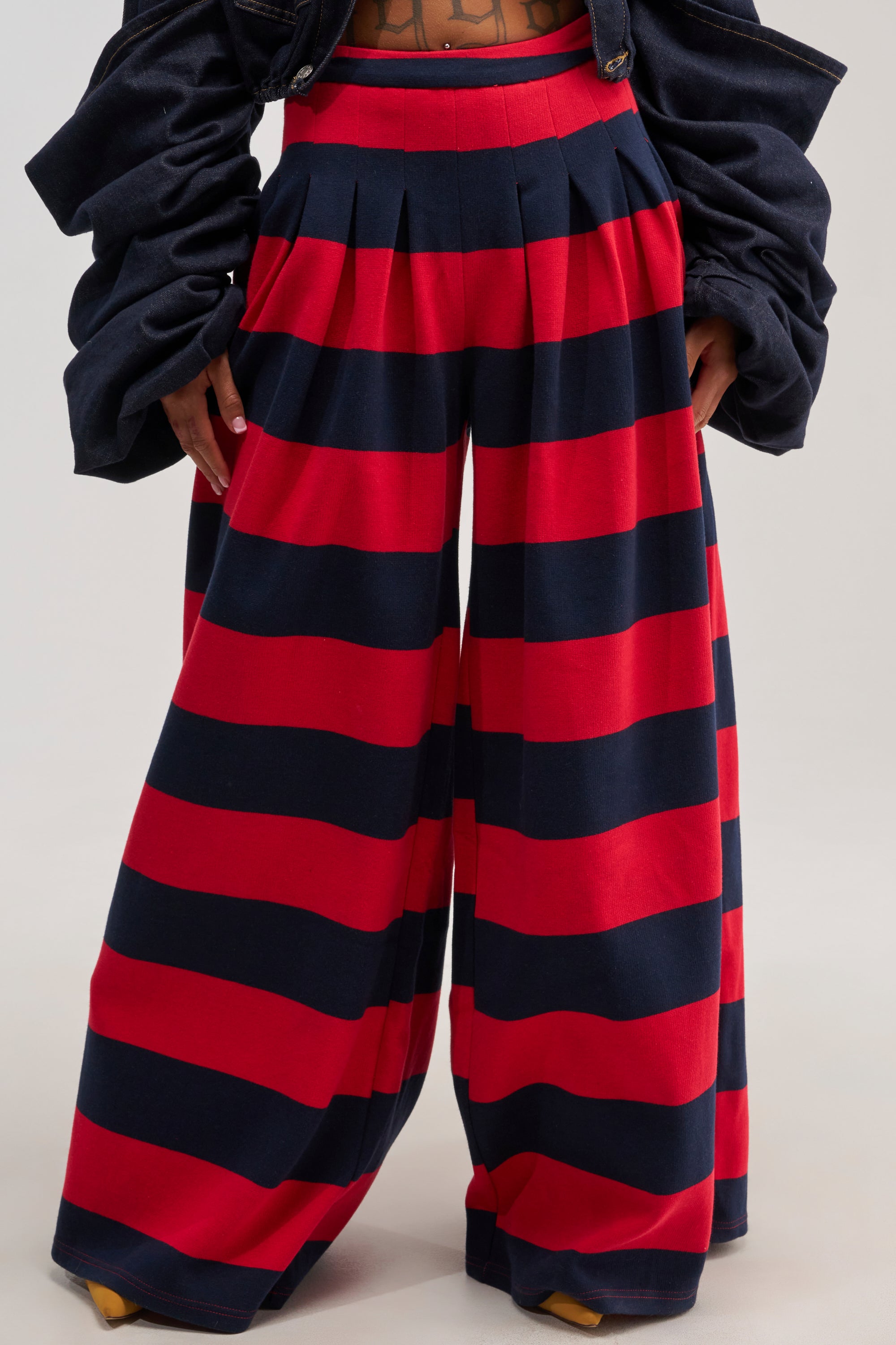 A person is seen wearing the SILVER COAST RED STRIPE TROUSER, featuring a high-rise fit and wide-leg silhouette with bold horizontal red and navy blue stripes, paired with a dark jacket. Only the lower torso and legs are visible.