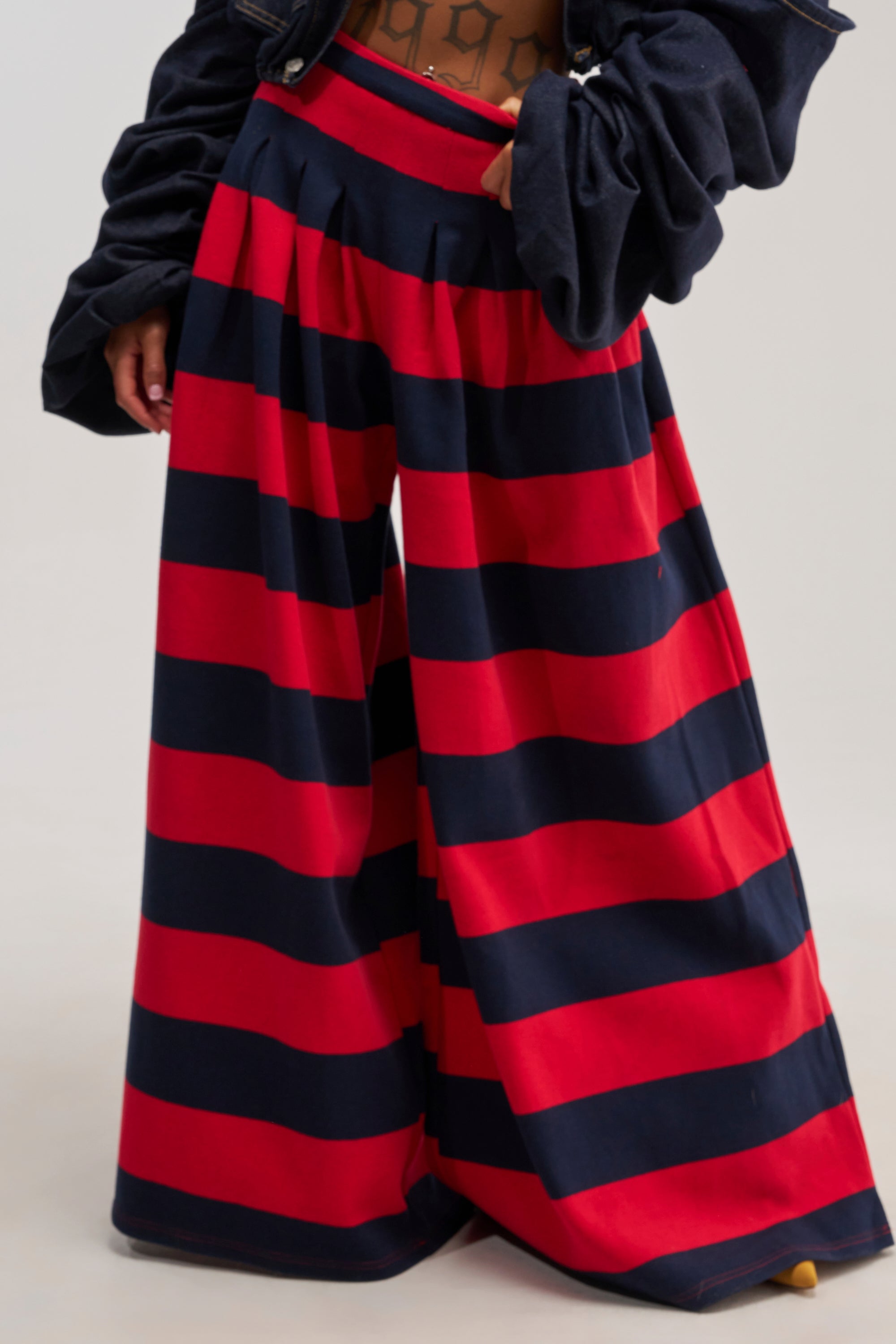 A person wears the SILVER COAST RED STRIPE TROUSER, which features a high-rise fit and a wide-leg silhouette. The trousers are loose-fitting with bold, horizontal red and navy blue stripes that extend to the floor. The individual's hands are partially visible against a plain, light-colored background.