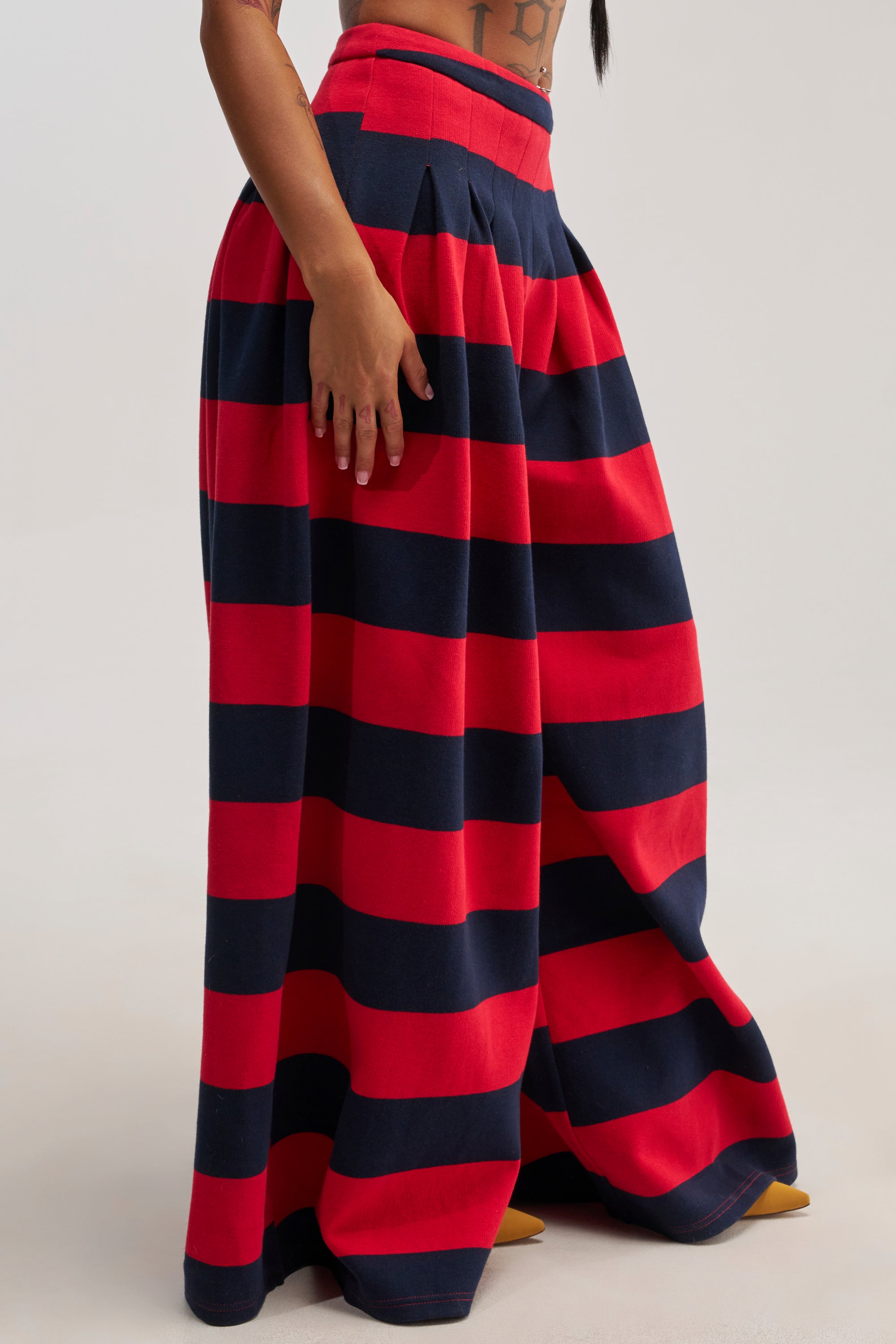 A person is wearing the SILVER COAST RED STRIPE TROUSER, which features a high-rise fit, red and navy blue stripes, and a wide-leg silhouette. Only the lower torso and upper legs are visible against a plain background.