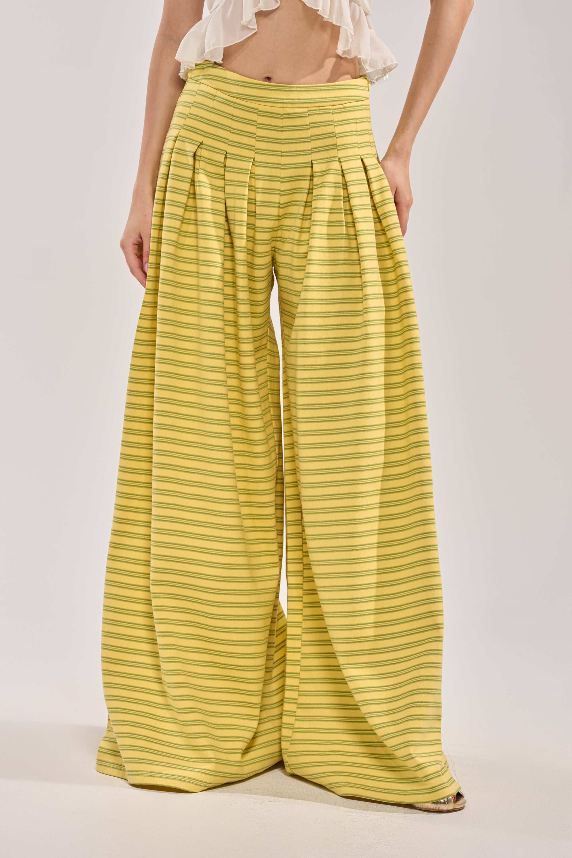 A person wears SILVER COAST YELLOW STRIPE TROUSER, which are high-waisted, wide-leg summer trousers with horizontal yellow stripes and pleats, paired with a ruffled white crop top against a plain, light background. 0