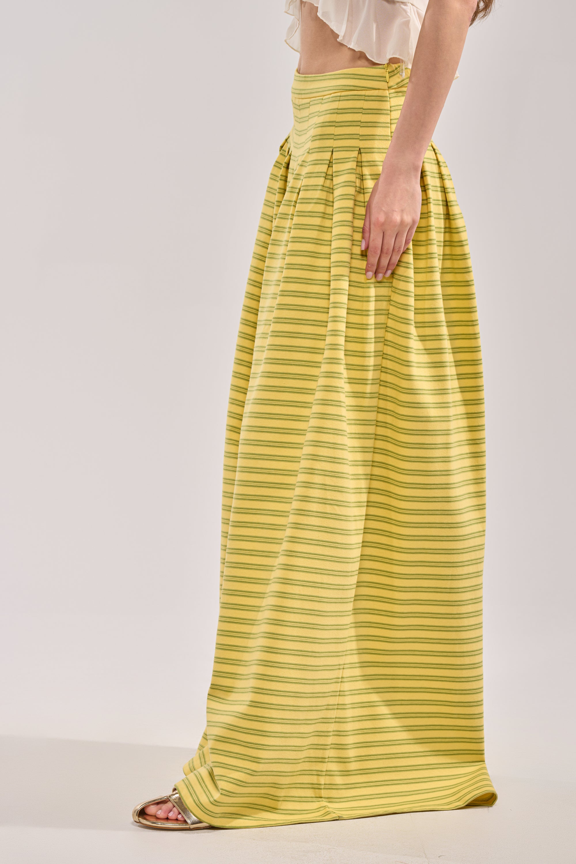 A woman models the SILVER COAST YELLOW STRIPE TROUSER, styled high-waisted as a pleated maxi skirt with a white cropped top and open-toe sandals; image is side view, face not visible. 1