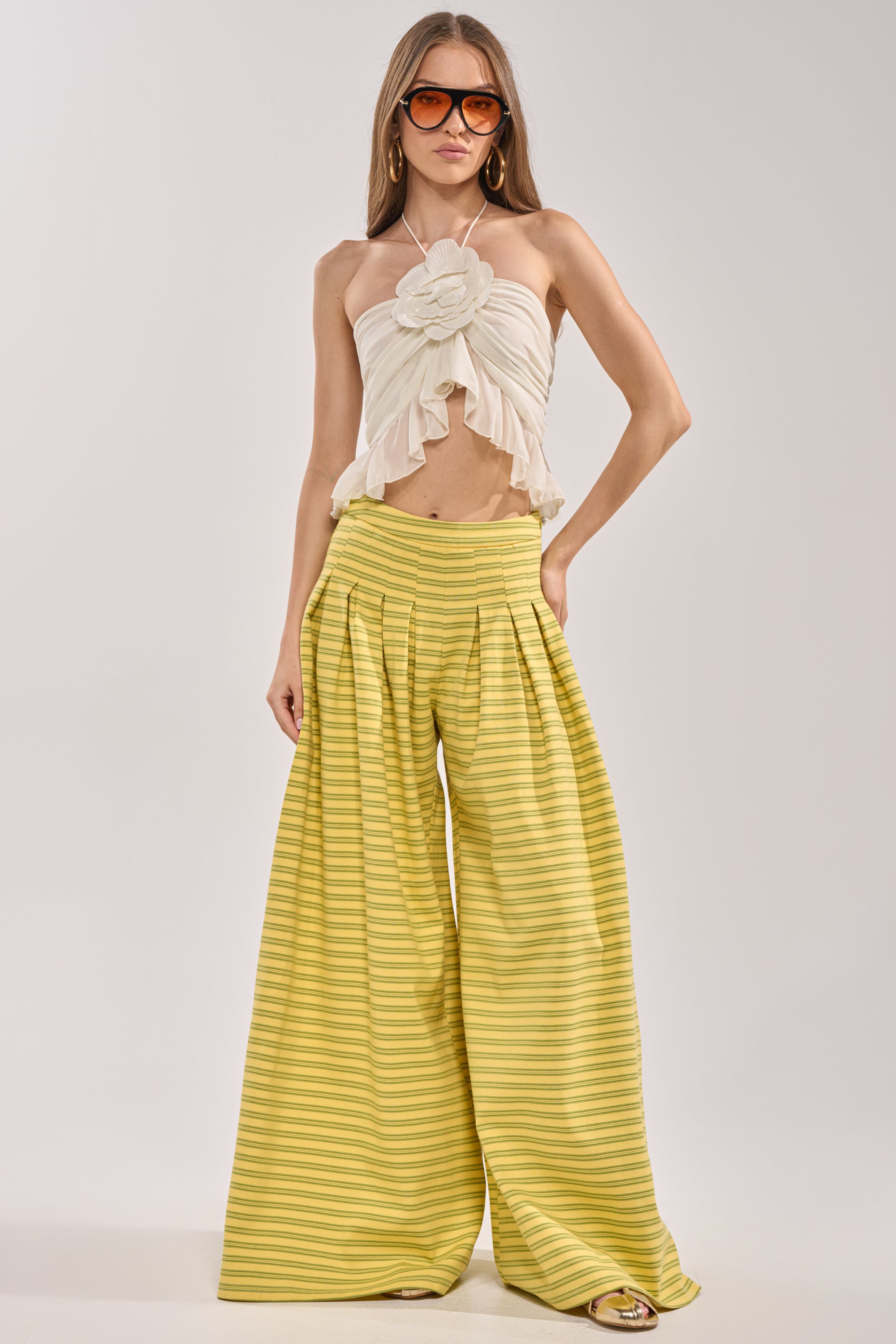 A woman models chic summer style in the SILVER COAST YELLOW STRIPE TROUSER, paired with a strapless cream ruffle top with a flower detail and oversized sunglasses, set against a plain background. 3