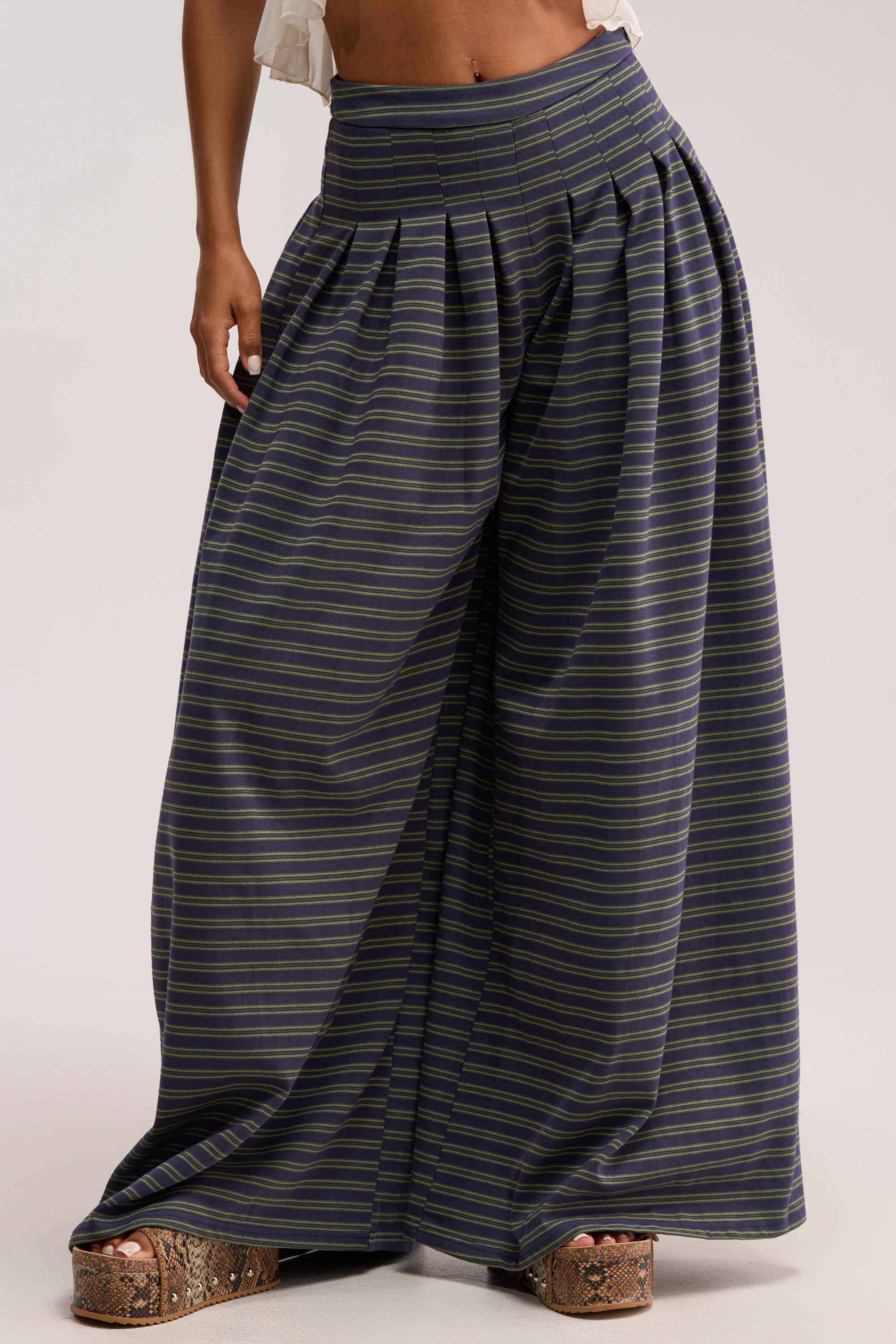 Wearing the GUESS WHO STRIPE PALAZZO PANT, featuring a high-rise fit, pleats, and wide-leg silhouette, paired with snakeskin-patterned platform sandals. Only the lower torso and legs are shown.