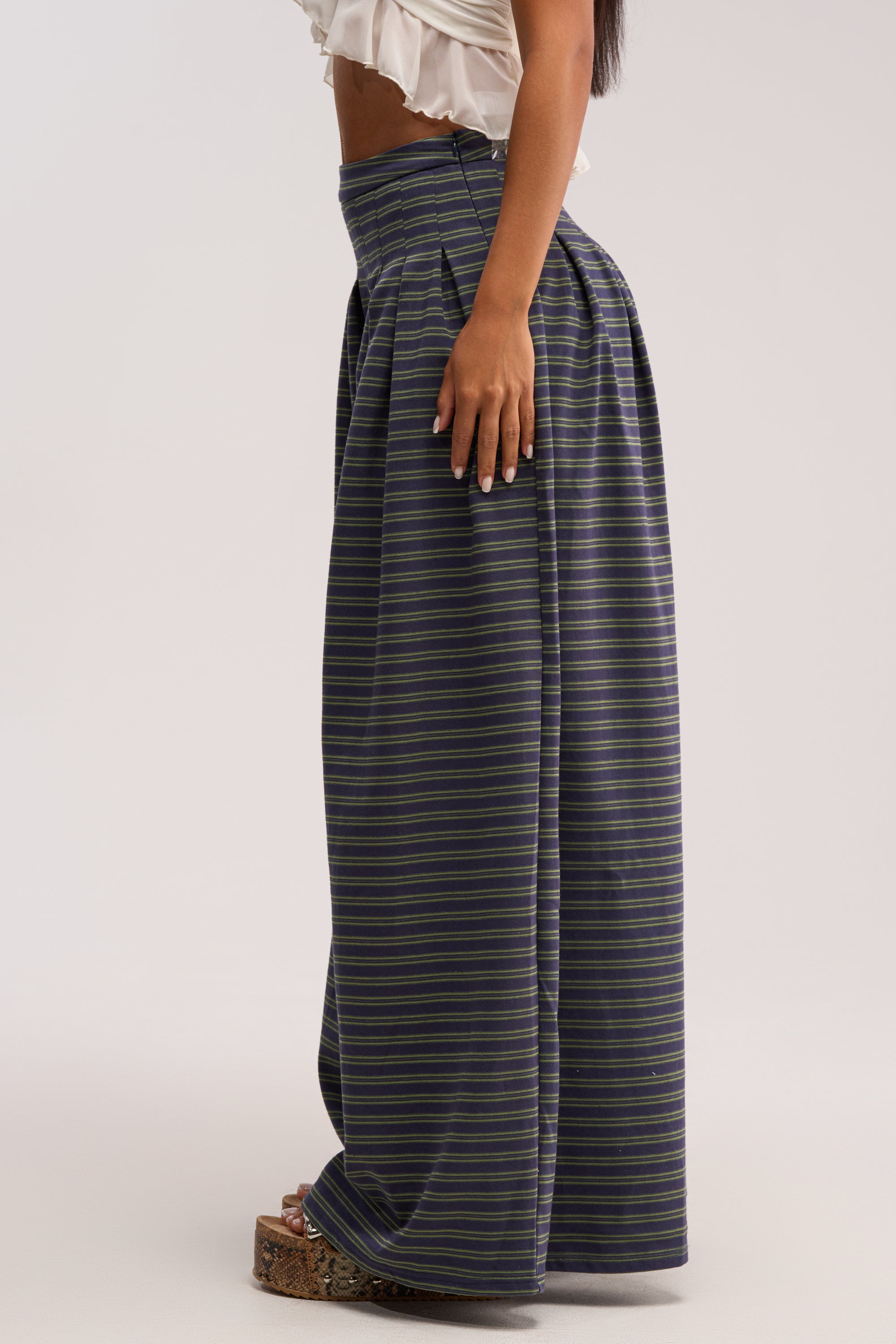 A person models the GUESS WHO STRIPE PALAZZO PANT, standing sideways to showcase its high-rise fit and wide-leg silhouette, styled with a white ruffled crop top and platform sandals for a relaxed look.