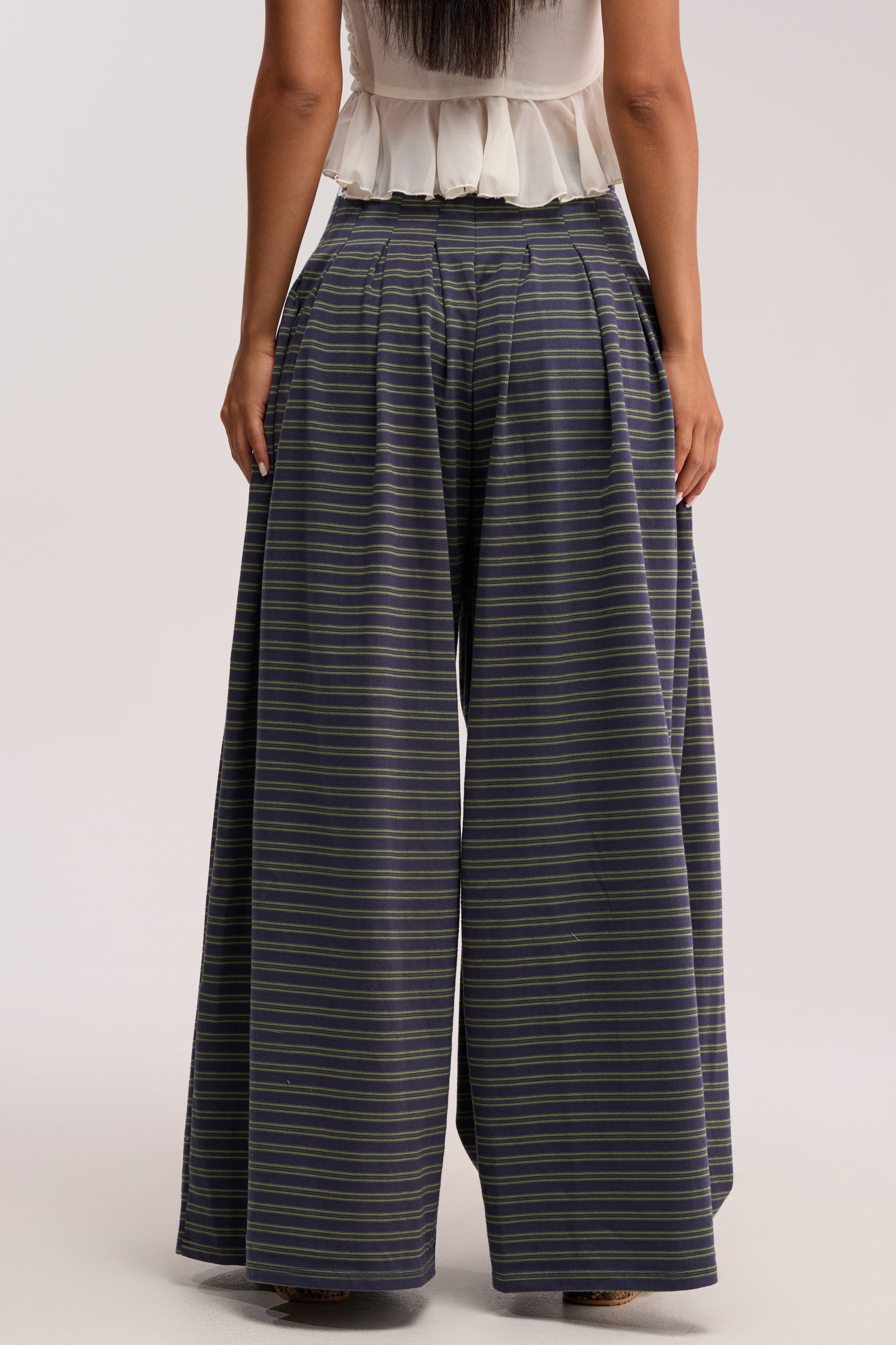 A person stands facing away, showcasing the wide-leg silhouette of the GUESS WHO STRIPE PALAZZO PANT in a high-rise fit with stripes, paired with a white sleeveless top against a plain background.