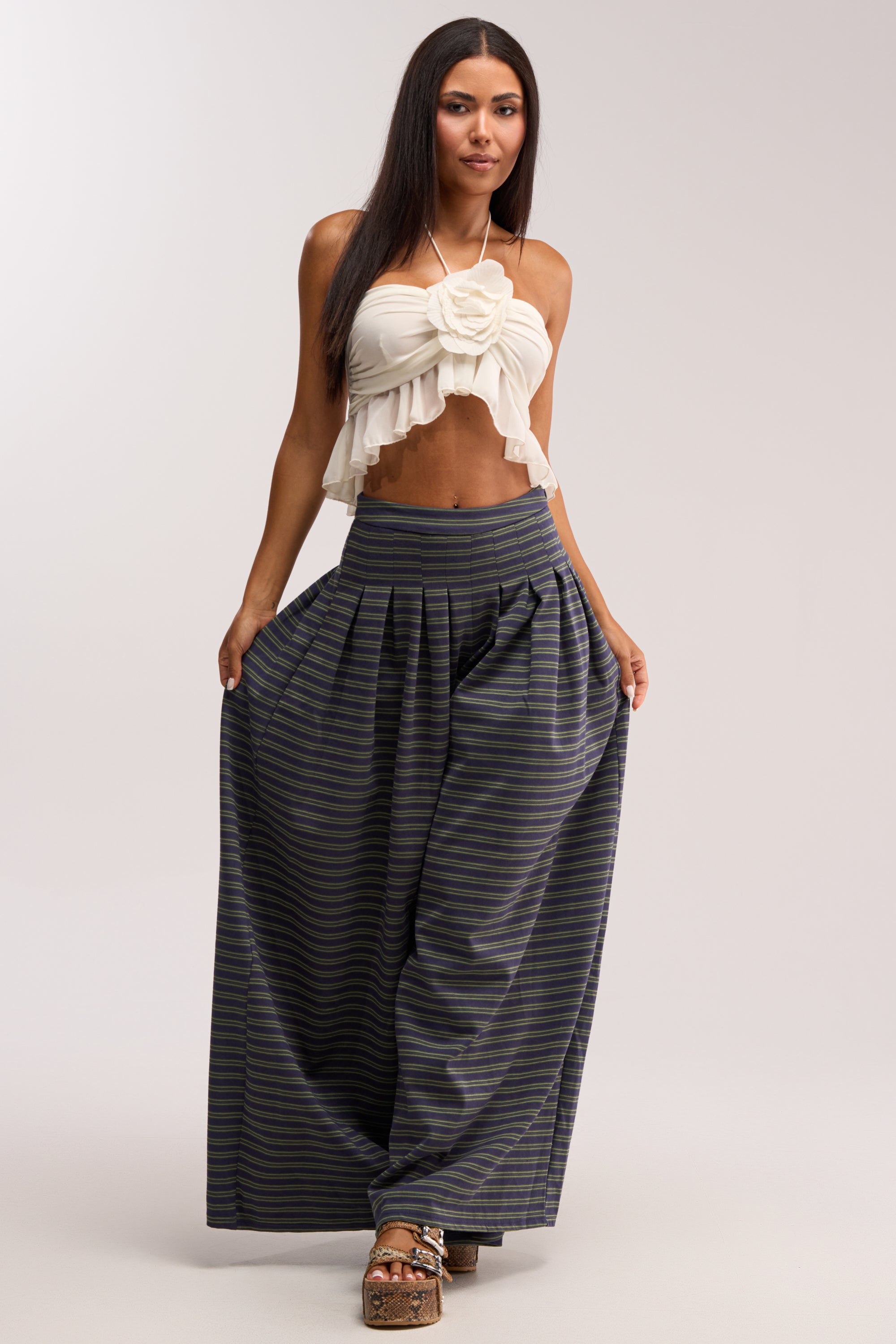 A woman with long dark hair stands against a plain background wearing GUESS WHO STRIPE PALAZZO PANT, featuring high-rise, wide-leg stripes, paired with a white ruffled halter top and platform sandals.
