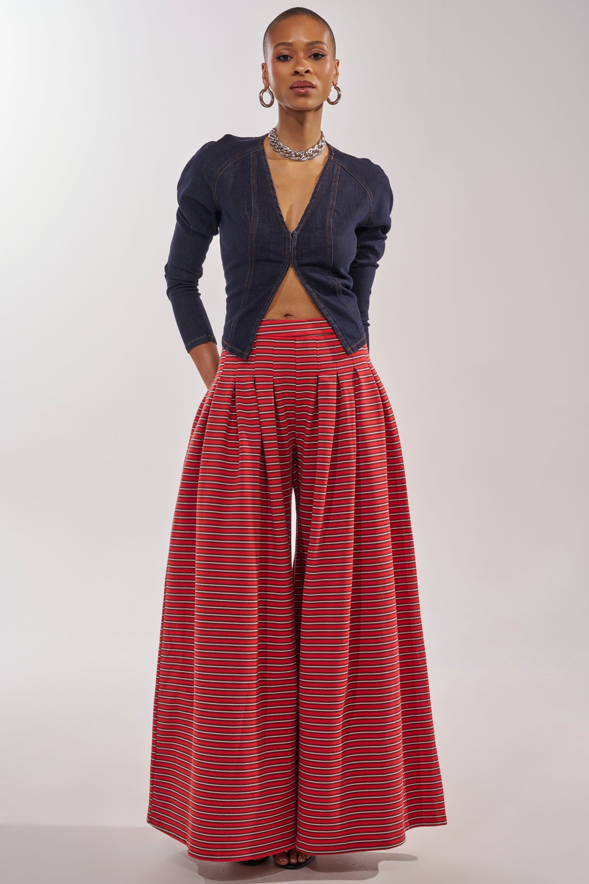 A person faces forward in a dark denim zip-up top with puffed shoulders and long sleeves, paired with SILVER COAST STRIPE TROUSER featuring a high-rise waist and colorful horizontal stripes. Hoop earrings and a necklace complete the look. 0
