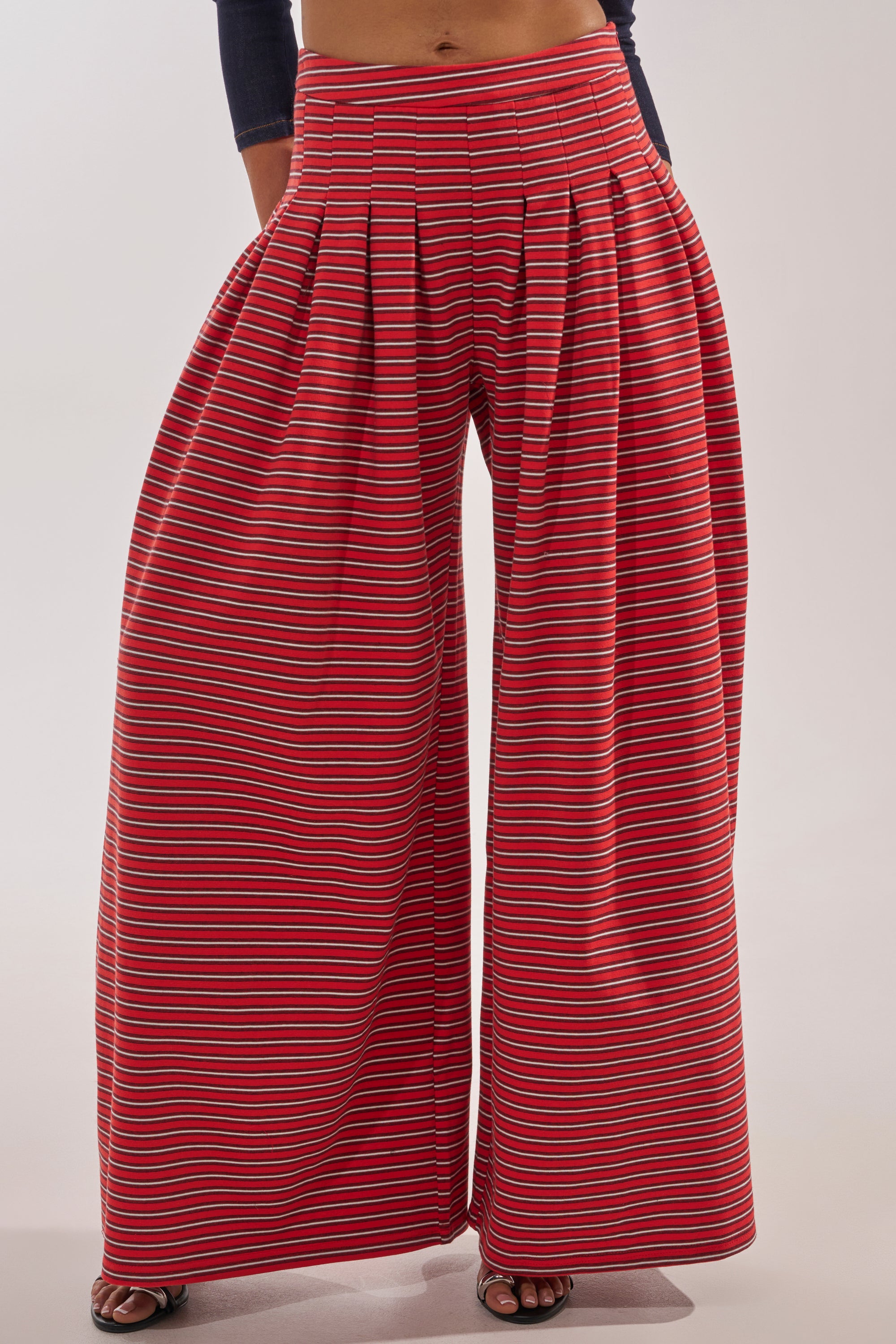 Model wearing the SILVER COAST STRIPE TROUSER—cotton wide-leg pants with a high-rise waistband and red and blue horizontal stripes, pleated at the waist. Only the lower body is visible against a plain background. 2