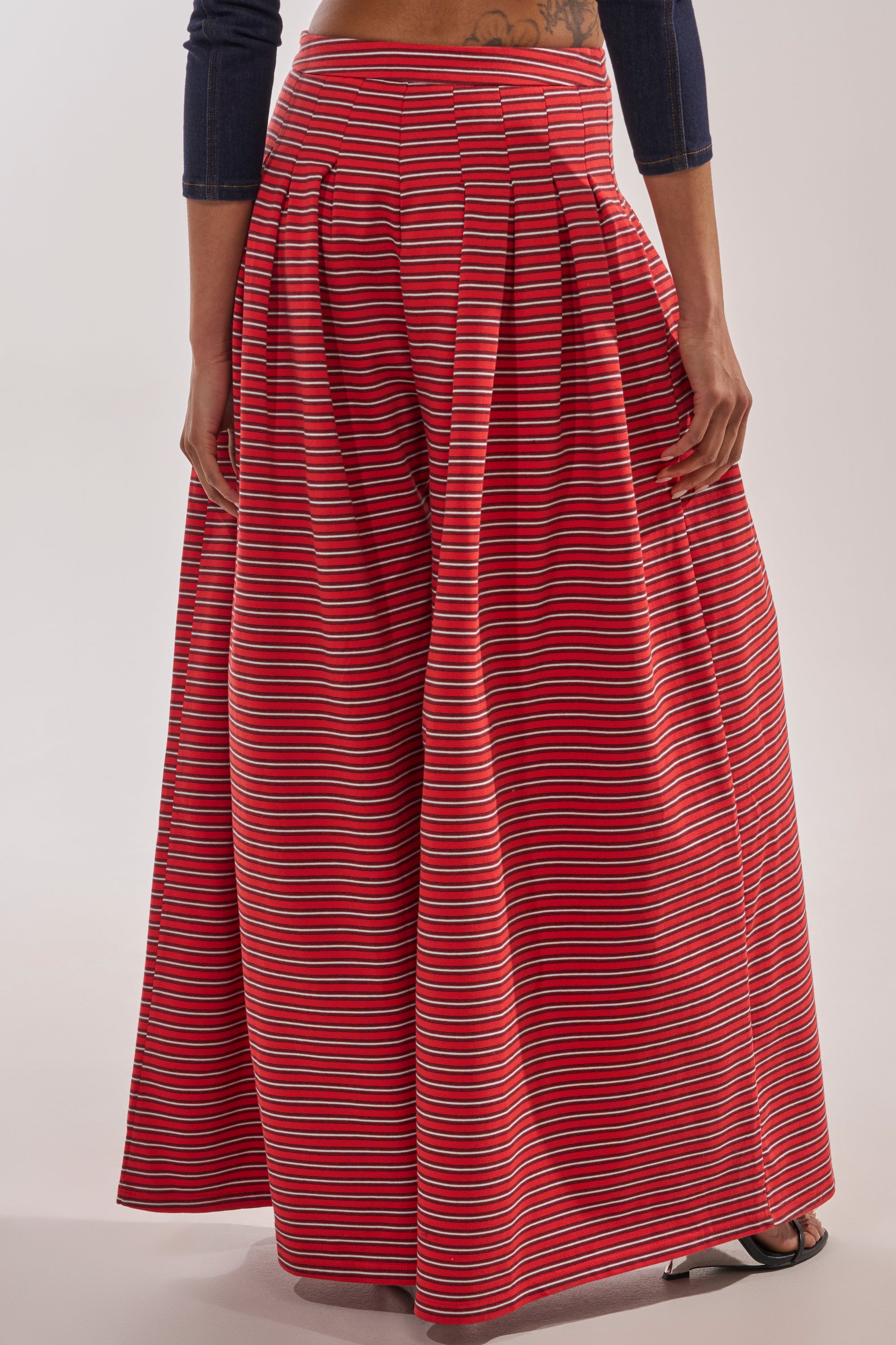 A person stands wearing the SILVER COAST STRIPE TROUSER—cotton, wide-leg pants with a high-rise waistband, red, white, and blue horizontal stripes, pleated waist, and pockets; their hands rest at their sides. 4