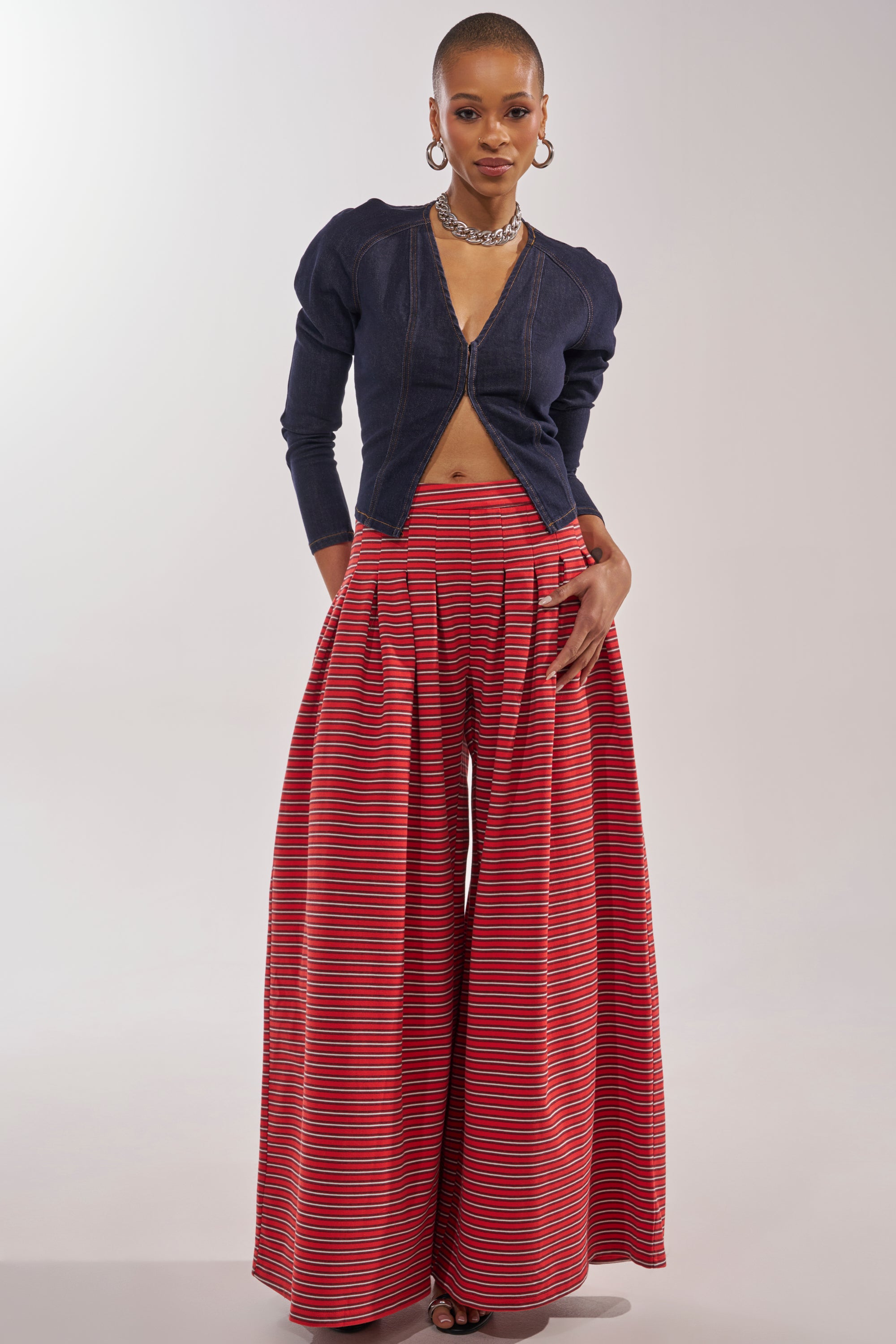 A person stands confidently in the fitted dark denim zip-up top, paired with SILVER COAST STRIPE TROUSER—high-rise, wide-leg cotton trousers in red with horizontal multicolored stripes—accessorized with hoop earrings and a choker. 5
